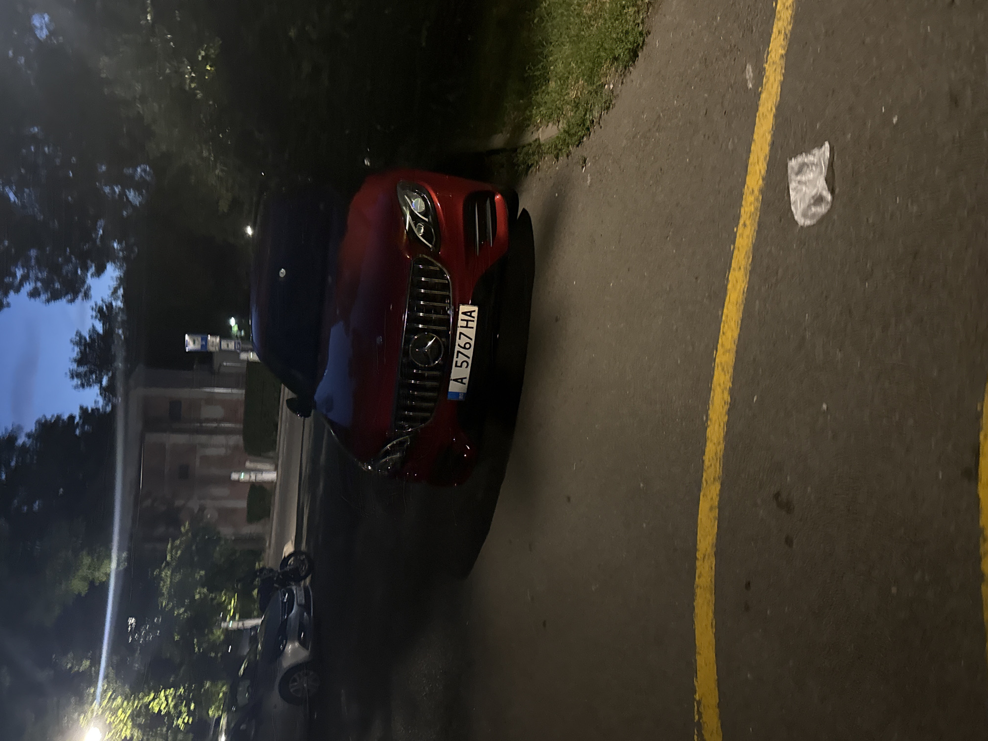 The image shows a street scene at dusk in Burgas, Bulgaria. A red Mercedes-Benz sedan is parked on the right side of the road, facing left. The car has a license plate that reads "A 5767 HA". In the background, there is a silver car and a motorcycle. Trees and a building are also visible. The road is paved and there is a yellow line painted on it. A crumpled plastic bag is lying on the road near the yellow line. The sky is dark blue, indicating that it is nighttime.
