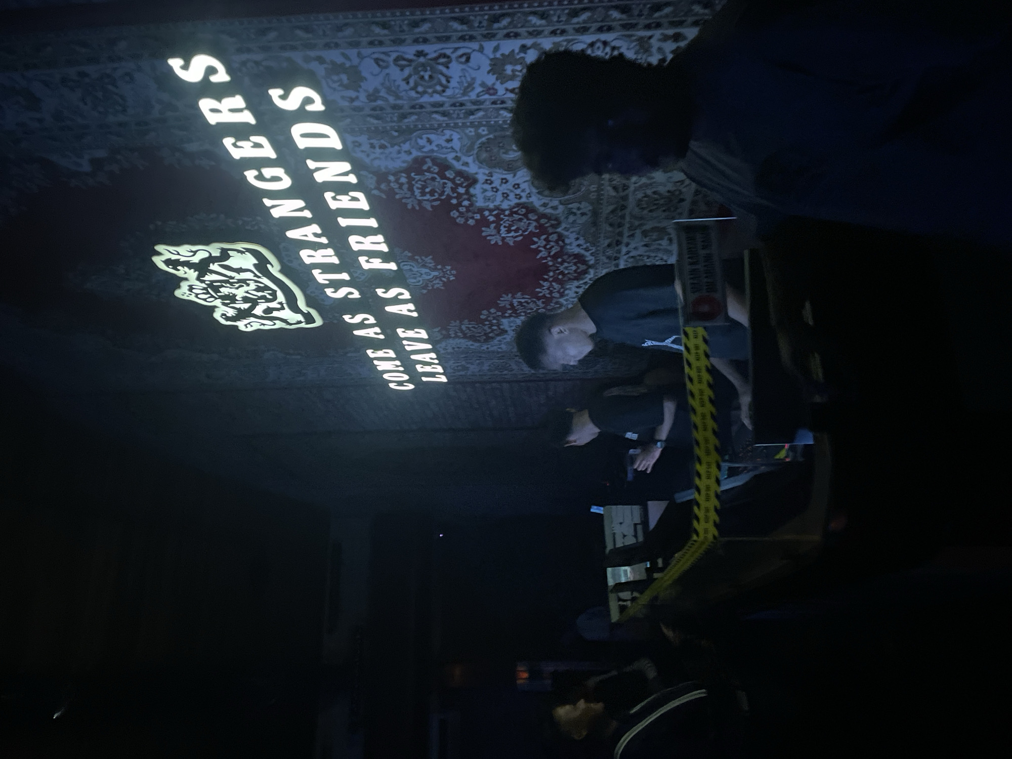 In a dimly lit indoor venue in North Jakarta, Indonesia, a group of individuals are gathered. A large, ornate tapestry covers the back wall, illuminated by a bright sign above it that reads "COME AS STRANGERS LEAVE AS FRIENDS" with a coat of arms emblem. In the foreground, several people appear to be working behind a barricade marked with yellow caution tape. One individual, facing away from the camera, is wearing a black t-shirt and appears to be holding something in their hands. To the left, another person in a black jacket looks towards the activity. The overall atmosphere suggests a social event or perhaps a gathering at a nightclub or bar.