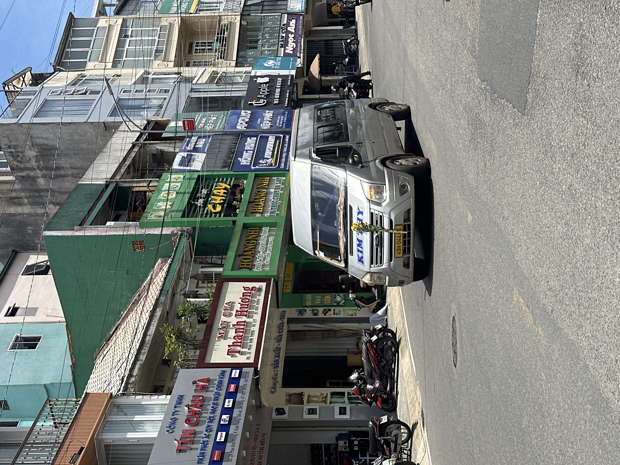 In the city of Da Lat, Vietnam, a silver Ford Transit van, identified by the license plate "49H-044.70", is driving down a paved street. The van has "KIM HY" written on its windshield. The street is lined with multi-story buildings housing various businesses. Among the visible signs are "TIN CHAU HA", "MAN CUA THANH HUONG", "HOANG NHI", "CHAY", "APOLLO", "Apple", and "NGOC ANH". Several motorcycles are parked on the sidewalk. A person wearing a dark helmet is riding a motorcycle. The sky is clear and blue. The buildings exhibit a range of architectural styles and colors, including teal, green, white, and beige. The environment appears to be an urban commercial area.