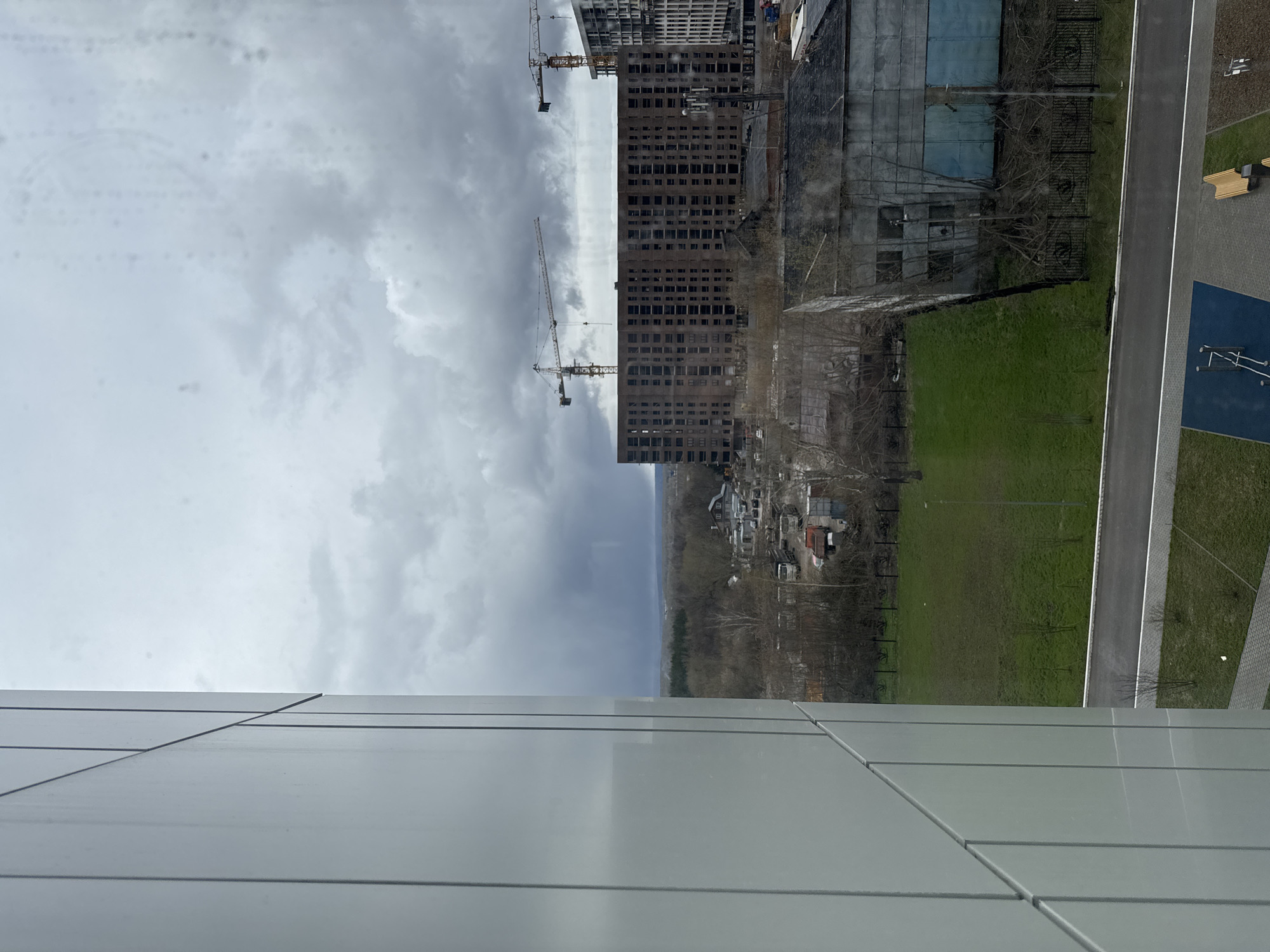 The image presents an outdoor scene viewed from an elevated perspective, encompassing urban development and natural landscapes under an overcast sky. In the immediate foreground, the light gray, paneled exterior of a modern building, from which the photograph was taken, occupies the left and bottom portions of the frame.
The midground is dominated by two large, multi-story buildings under construction, featuring brick and concrete skeletal structures with numerous window openings. Two tall tower cranes are prominent beside these buildings, indicating ongoing development. To the right, a paved road or pathway runs diagonally, separating sections of vibrant green grass. Adjacent to the path is a children's play area with a blue surface and white structures, and a wooden bench. Further back, another green field with bare trees and a metal fence borders an older, possibly industrial-style building with a partially blue facade and visible windows.
In the background, a dense line of deciduous trees, mostly bare, forms a forest, behind which a cluster of smaller buildings or houses is visible at the horizon. The sky is uniformly cloudy and overcast, creating a soft, diffused light across the scene. The overall impression is one of a developing area, blending new construction with existing infrastructure and natural elements. No distinct text is visible in the image. The scene depicts a daytime view in what appears to be a suburban or semi-rural setting, consistent with the location context of Borovoye Matyushino, Russia.