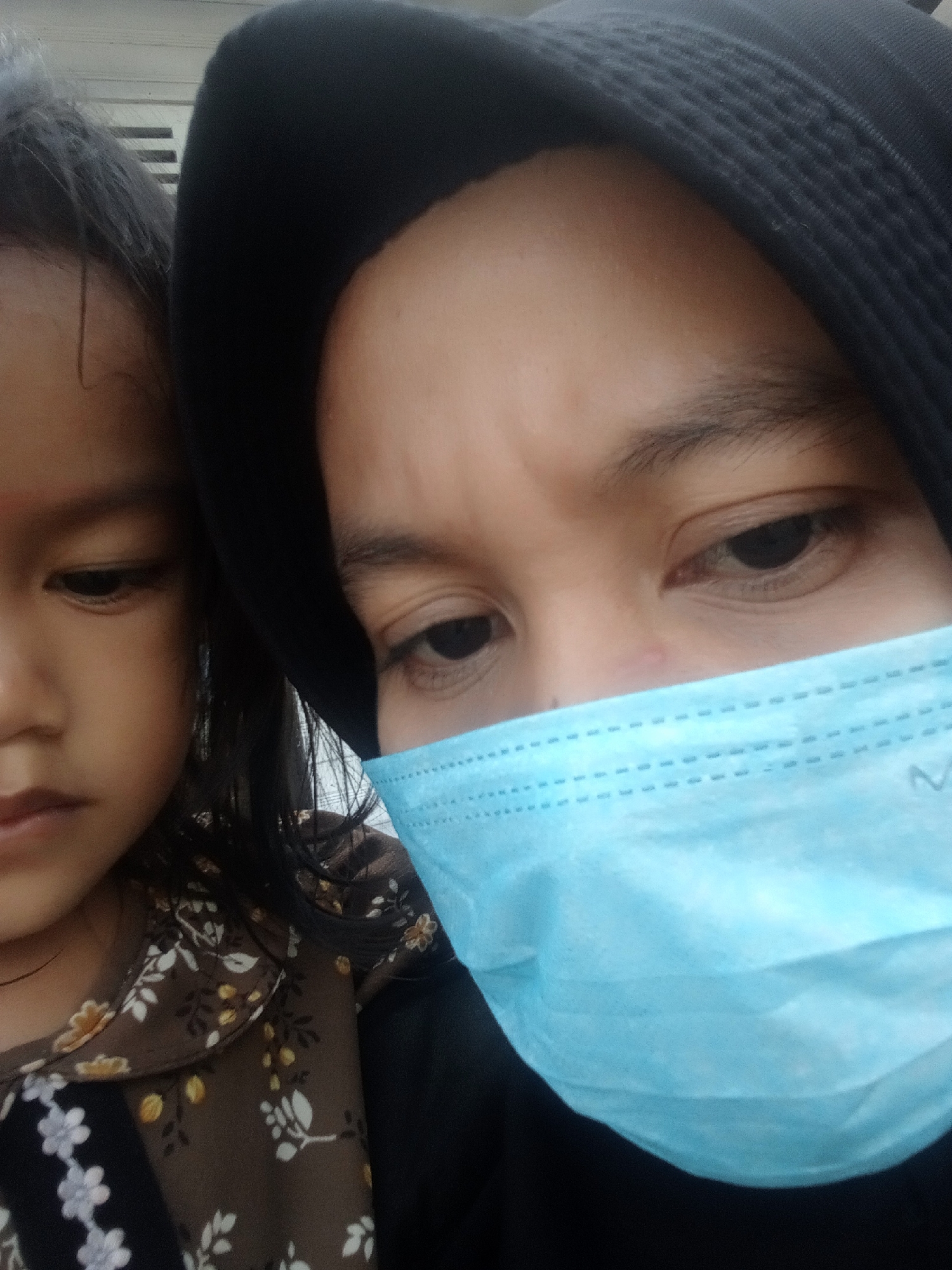 The image shows a close-up of two individuals, likely a mother and child, wearing black hijabs. The person in the foreground, appearing to be an adult, is wearing a light blue surgical mask that covers their nose and mouth. Their eyes are visible, looking towards the camera. To the left of this person, a child is partially visible, with their face also in profile. The child has dark hair and is wearing a brown shirt with a floral pattern. The background is indistinct, but appears to be an indoor or shaded outdoor setting with horizontal blinds visible in the upper left corner. There are no specific visual cues in the image that indicate the location is the city of Campago, Indonesia.