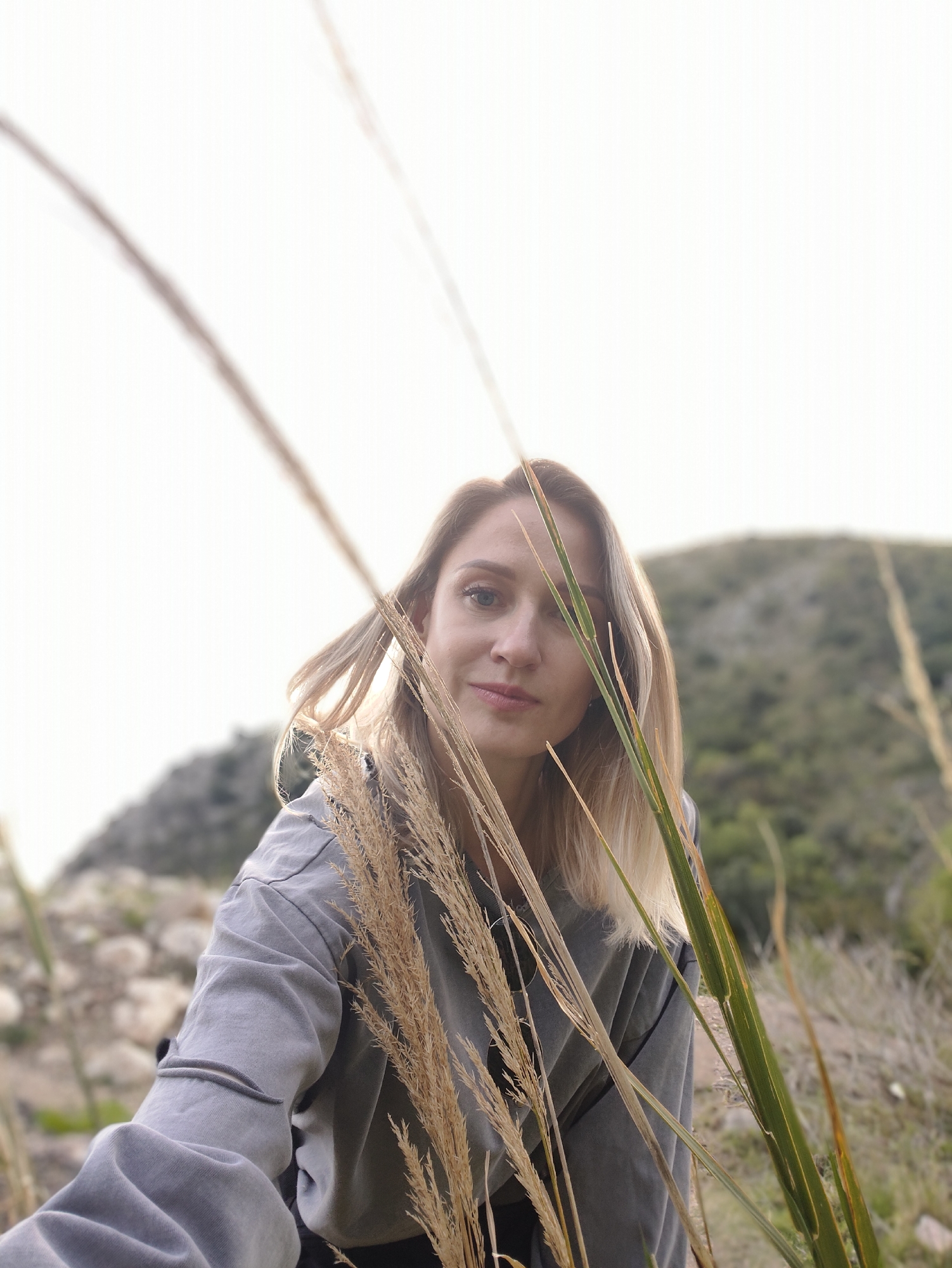 A fair-skinned woman with blonde hair is the primary subject of the image. She is wearing a gray sweatshirt or jacket. The woman is positioned in the foreground, surrounded by tall grass or reeds. A small mountain is in the background. The location is Rafailovići, Montenegro.