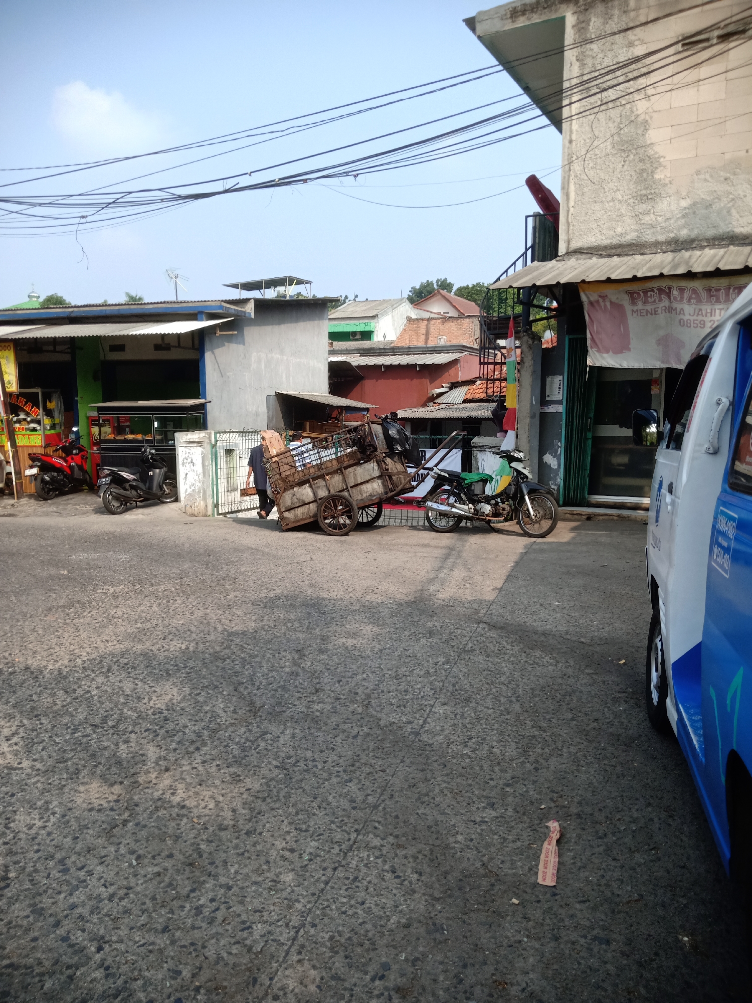 A daytime, medium-shot, outdoor, eye-level, full-color, vertical-format photograph depicts a street scene in East Jakarta, Indonesia. The foreground is dominated by a rough asphalt street, casting long shadows. A damaged cart laden with various items, including boxes and bags, is parked on the street, partially obscuring a metal gate. Two individuals, one wearing a blue shirt and the other a darker garment, are near the cart.
To the left, two motorcycles are parked. The front motorcycle is red and black, and the one behind it is black. Behind the motorcycles are storefronts with awnings. The storefront on the far left has a yellow sign with red lettering. Next to it, a green storefront features a display of food.
In the middle ground, a white and green motorcycle is parked near the metal gate. The gate opens onto a narrow pathway with buildings beyond. A white van with blue detailing is parked on the right side of the street, facing away from the viewer.
In the background, a variety of buildings and structures are visible, including a mosque with a green dome on the far left. Power lines crisscross the sky. The sky is clear and blue with a few white clouds. The overall impression is one of a busy, urban street scene with commercial activity.