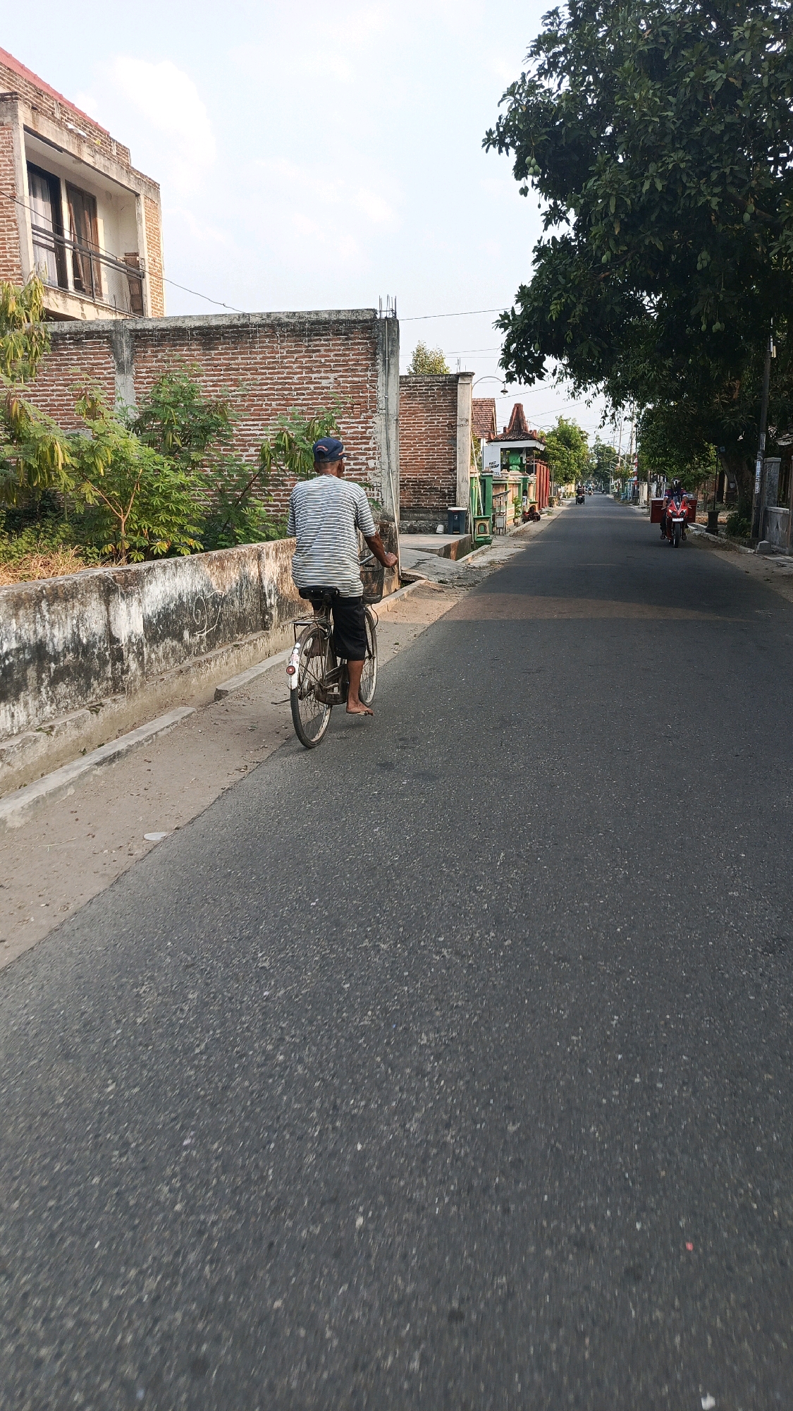 A man in a striped t-shirt and dark shorts rides a bicycle down a paved road in Cekok, Indonesia. He wears a blue baseball cap and appears to be barefoot. The bicycle has a basket on the handlebars and a fender with a reflector on the rear wheel. The road is lined with buildings and trees. On the left, a low, weathered concrete wall separates the road from a brick wall and a two-story building with an unfinished facade. Green foliage grows along the wall. On the right side of the road, there are more buildings and trees, including a large tree with green leaves. In the distance, two people on a red scooter are also traveling down the road. The sky is clear with a few clouds. The overall impression is a sunny day in a rural or suburban area.