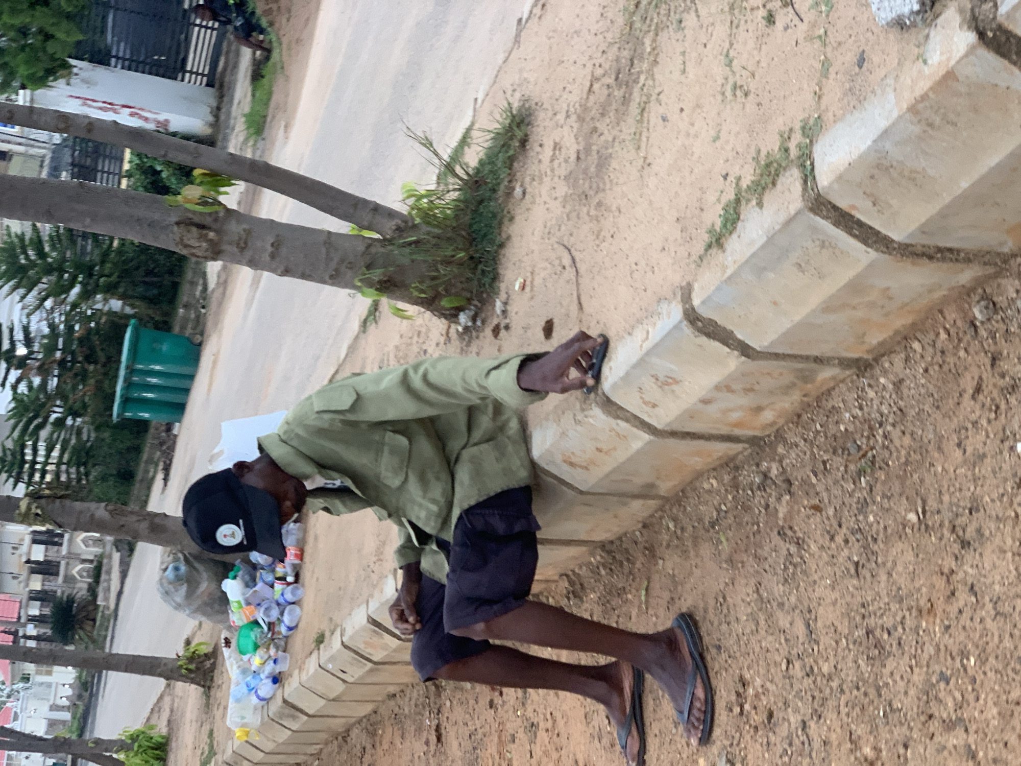 A man wearing a green jacket, black shorts, and a black baseball cap sits on a concrete curb next to a pile of plastic bottles. He is looking down at a dark-colored object in his right hand. His left hand rests on his left knee. The ground beside the curb is covered in dirt and small stones, with some patches of dry grass. Behind him, a paved road is visible with trees and buildings in the background. Two green trash bins are present further down the road. The scene appears to be an outdoor area in Abuja, Nigeria, possibly a roadside or a public space.