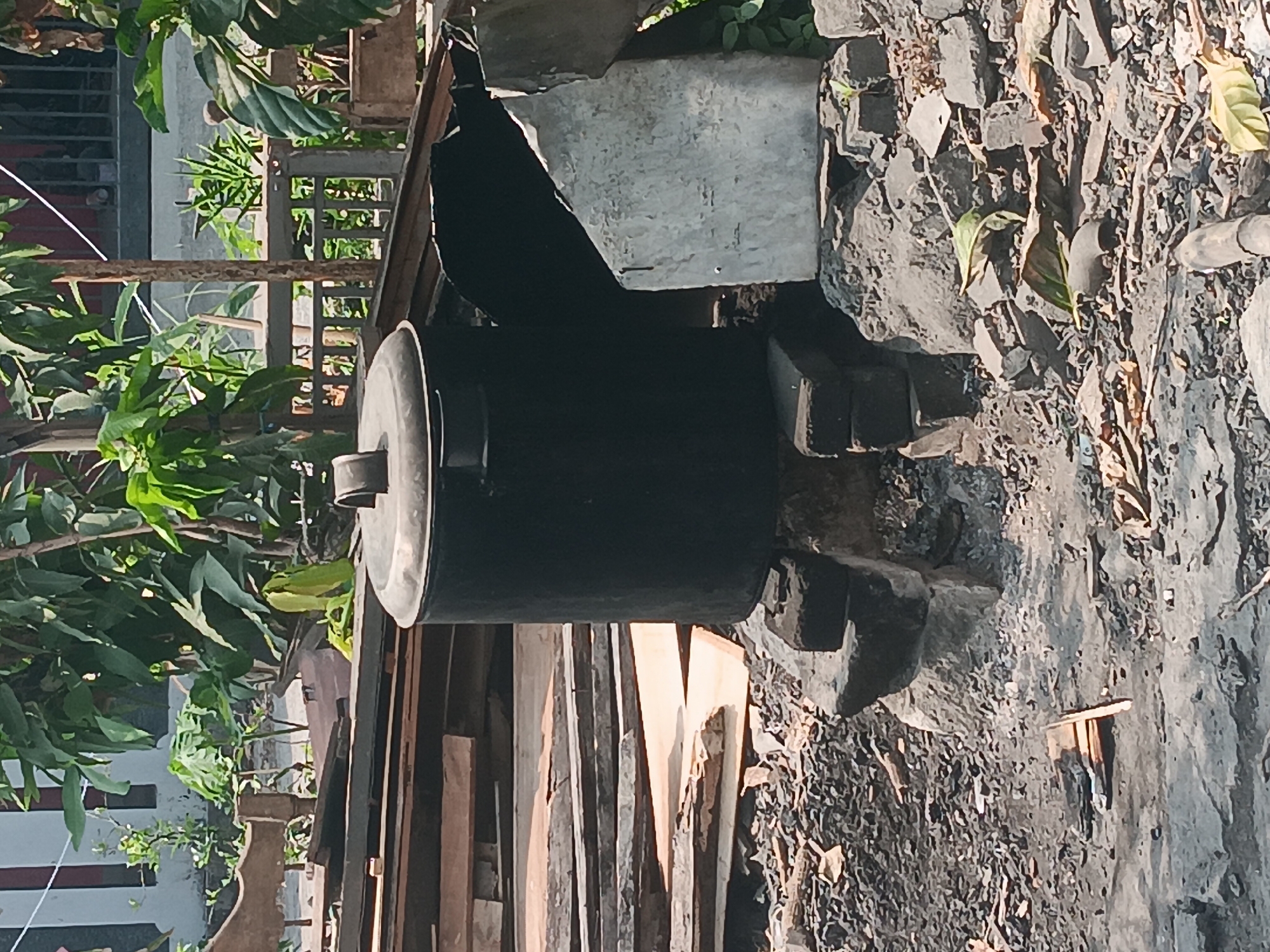 In Batursari, Indonesia, a black metal cauldron sits on a makeshift stand of bricks and stones. The cauldron is situated outdoors, surrounded by debris. To the left of the cauldron, a stack of wooden planks is visible. Behind the cauldron, dense green foliage provides a natural backdrop. The ground is covered in a mixture of dirt, small stones, and scattered leaves, indicating an unkempt outdoor area. A large, broken concrete slab leans against the right side of the cauldron. The scene suggests a functional, perhaps rustic, setup for cooking or processing materials.