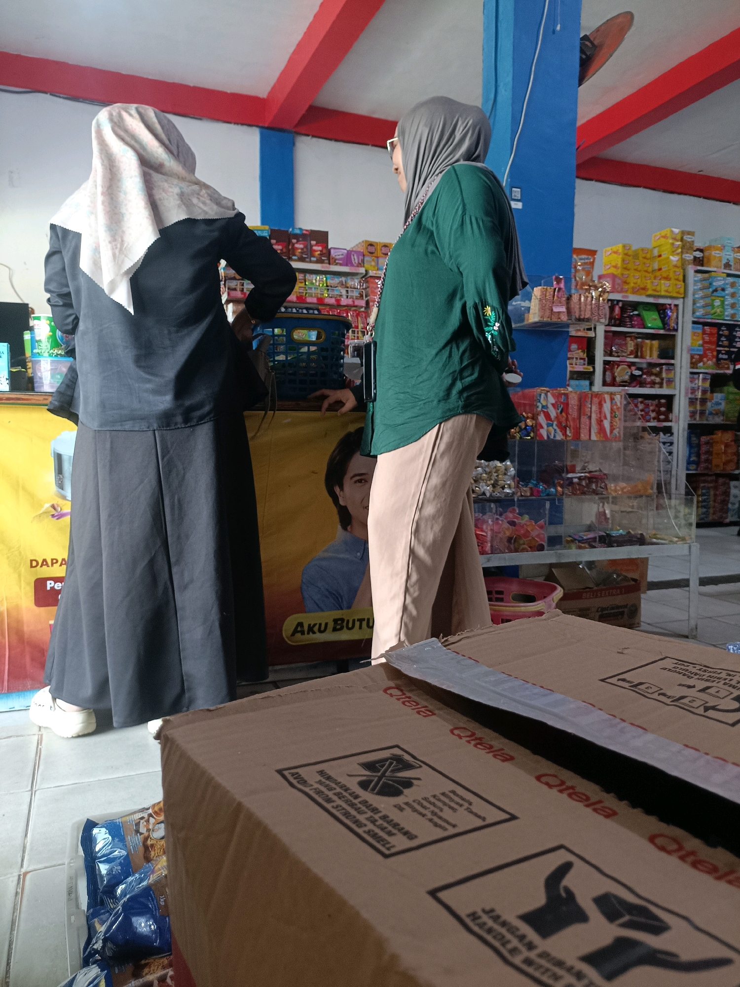The image shows the interior of a small shop or convenience store, possibly located in the city of Palopo, Indonesia. Two women are the primary subjects. The woman on the left, seen from the back, wears a black abaya and a light-colored patterned hijab. She has white sneakers on her feet. The woman on the right, facing left, is wearing a dark green long-sleeved top with embroidered details on the sleeves and light brown trousers. She also wears a grey hijab and glasses.
The shop displays shelves stocked with various packaged goods, including snacks and drinks. A clear plastic display case filled with candies is also visible. In the foreground, two cardboard boxes are stacked, one obscuring part of the other. The boxes have visible markings and text, including the word "Qtela" and instructions in a foreign language. A yellow banner with a picture of a man and text in Indonesian, "Aku Butu", is partially visible in the background. The floor appears to be tiled. The ceiling has red beams and blue sections.