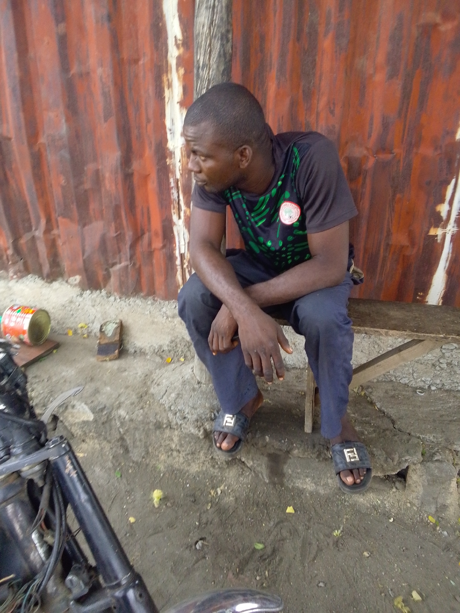 A man sits on a bench in front of a rusty corrugated metal wall, likely in Lokoja, Nigeria, based on context. He wears a t-shirt with a circular patch and dark trousers, along with black sandals with a white square design. A motorcycle is partially visible in the foreground. A discarded can and a small, brick-like object are on the ground near the bench. The ground is uneven, with a mix of dirt and concrete.
