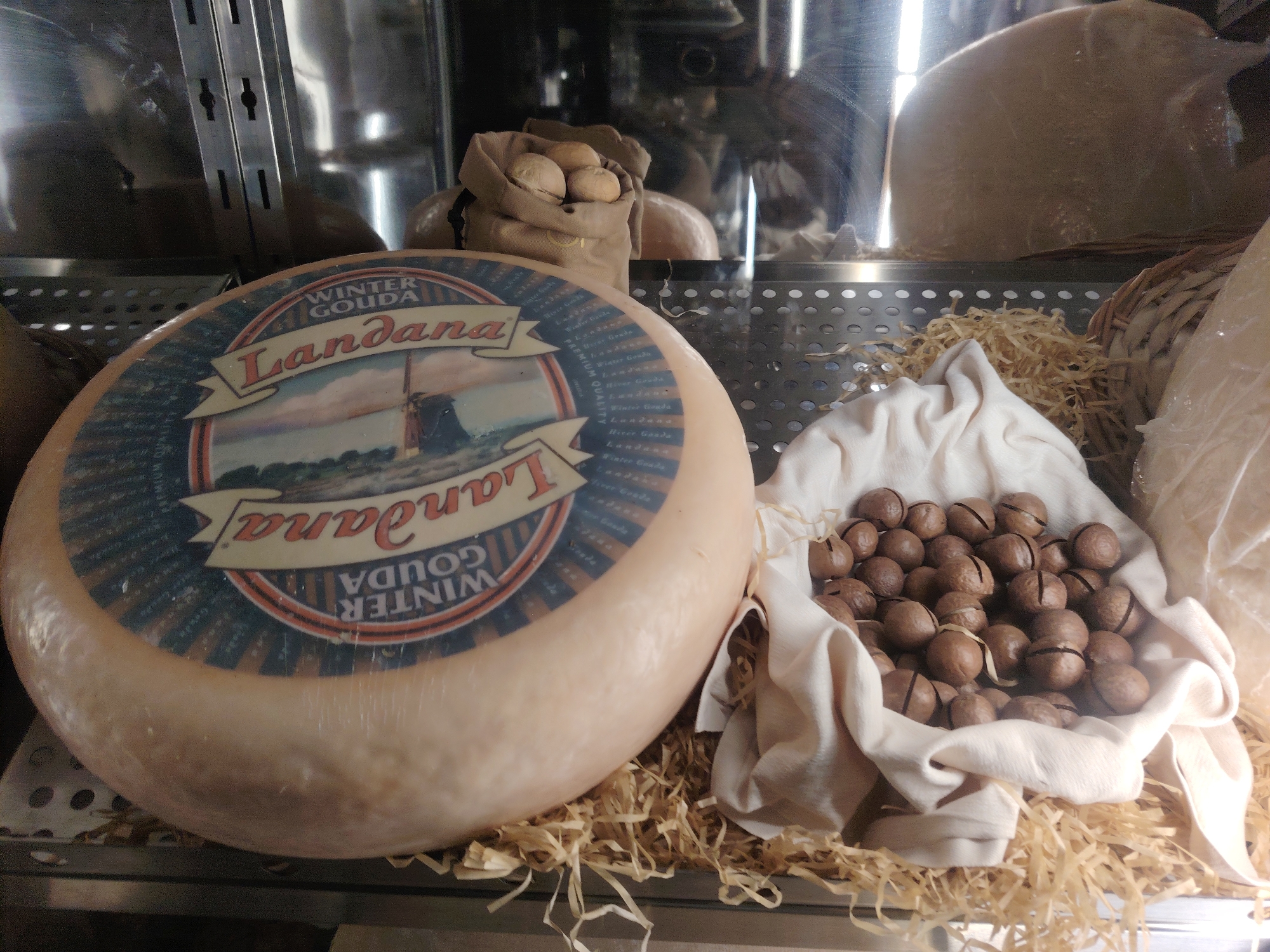 A large, round wheel of cheese, possibly Gouda, with a label indicating "Winter Gouda Landana," is displayed on a bed of straw. To the right of the cheese, a small cloth bag filled with nuts, possibly macadamia nuts, is visible. The cheese and nuts are presented on a shelf in what appears to be a retail setting, possibly a specialty food store or market, within the city of Konigil, Uzbekistan. The background shows metallic shelving and reflections, suggesting it is inside a display case or refrigerated unit.
