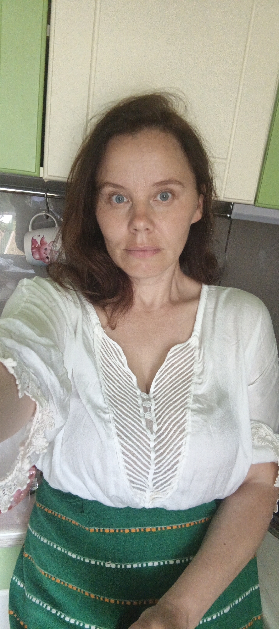 A woman with brown hair and blue eyes is standing in a kitchen, wearing a white blouse and a green skirt. The blouse has lace trim on the sleeves and a V-neckline with sheer detailing. The skirt is a dark green with a patterned border of white and orange dots. Behind her, there are cabinets, a mug hanging on a hook, and what appears to be a refrigerator.
