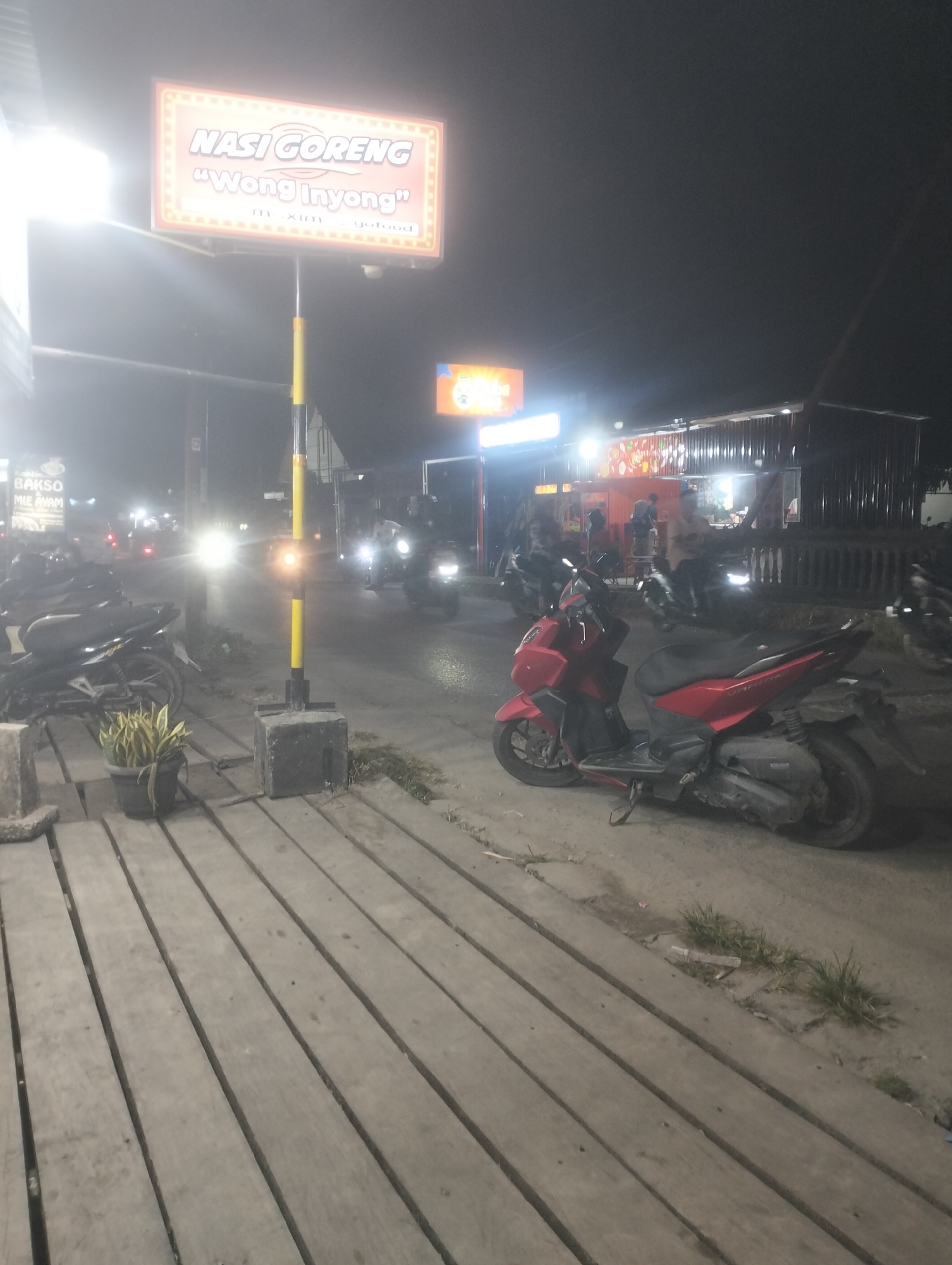 The image depicts a nighttime street scene in the city of Dumai, Indonesia, likely in a commercial area. A large, illuminated sign at the top left of the frame reads "NASI GORENG 'Wong Inyong'" in orange and white lettering, indicating a fried rice establishment. Below this, a yellow and black striped pole stands prominently.
In the foreground, a wooden walkway is visible, with a potted plant on its left. To the right of the walkway, a red scooter is parked. Several other motorcycles are also present in the scene, some with their headlights on. The background shows a row of food stalls or small shops, with a corrugated metal roof structure. People are visible in the distance, near the food stalls, and a few vehicles are on the road. The overall atmosphere is that of a bustling street food area at night.