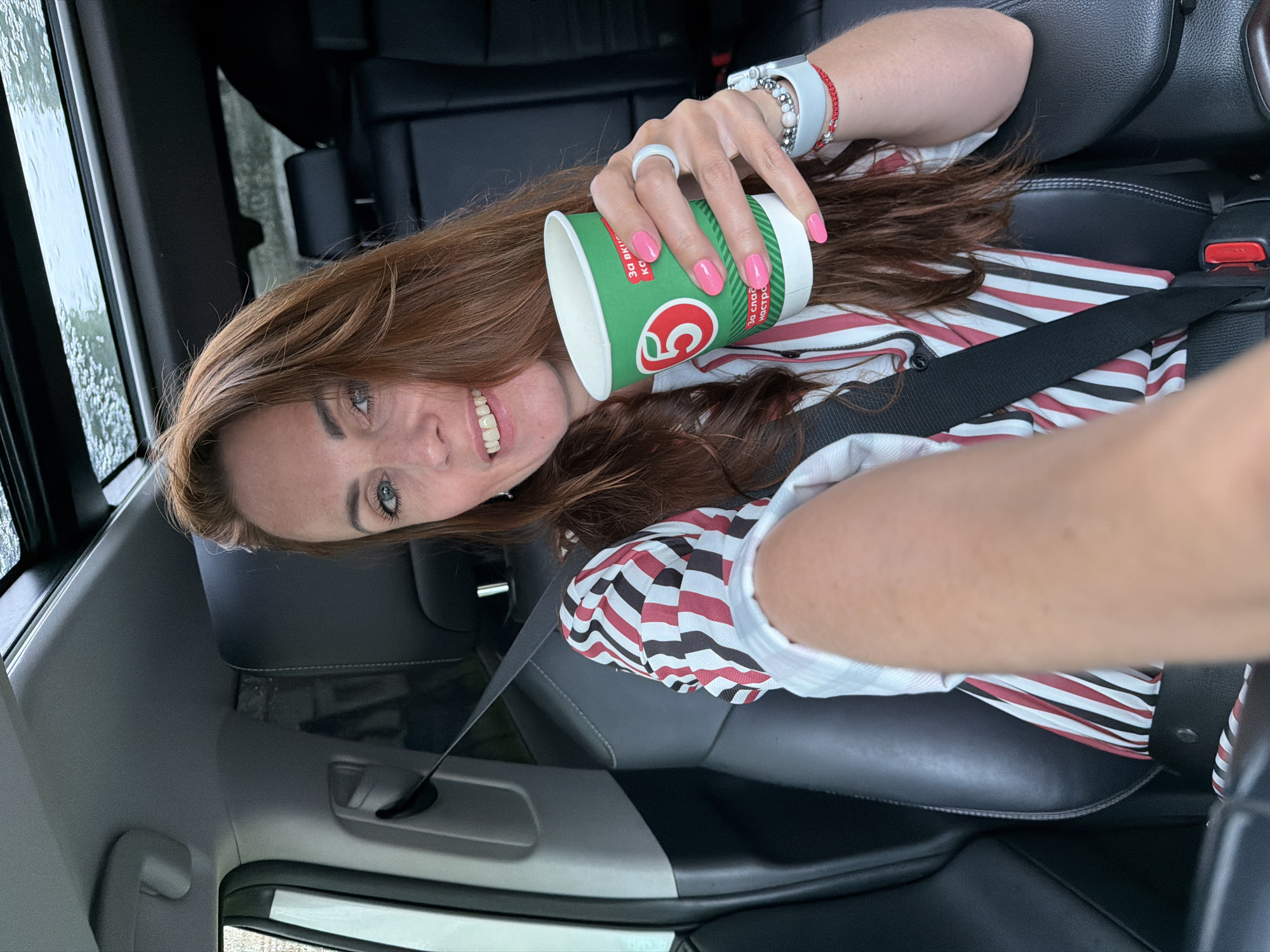 An adult woman with long brown hair and blue eyes is seated in the driver's seat of a car, holding a green and white paper cup with a red '5' logo. She is wearing a striped shirt and has bright pink nail polish. The car has black leather seats and a sunroof through which a cloudy sky is visible. The woman is looking at the camera and smiling, with her right arm extended as if taking a selfie. A seatbelt is visible across her body. The scene appears to be taking place in Birobidzhan, Russia.