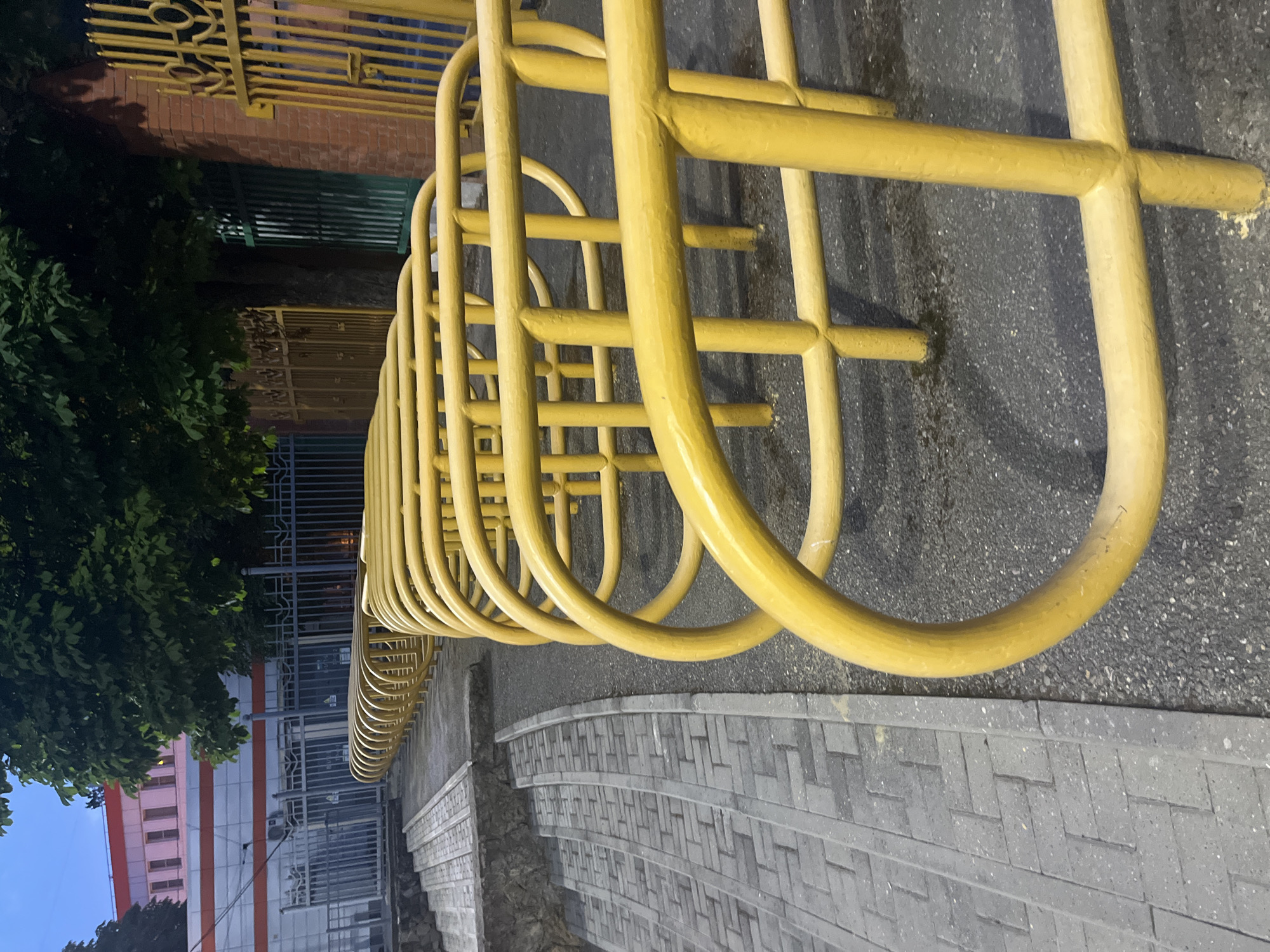 This is a photograph of an outdoor scene, likely in Krasnodar, Russia, taken during the evening or twilight hours. The primary subject is a long, curved yellow metal bicycle rack. The rack is constructed from thick, rounded tubes and extends horizontally across the frame, creating a repeating pattern of arches. It is positioned on an asphalt surface.
To the left of the bicycle rack, a decorative yellow metal fence and a brick wall are visible. Further to the left, dark green foliage from a tree obscures part of the scene. Behind the tree, a building with a light-colored facade and orange stripes can be seen. Another section of a building with a pale pink and white facade is also partially visible. A dark metal fence with intricate patterns runs along the edge of the property, separating it from the street.
In the foreground, a paved walkway made of gray rectangular stones laid in a herringbone pattern curves gently away from the bicycle rack. The lighting suggests it is either dusk or a street-lit area, with some shadows cast by the bicycle rack. There are no people visible in the image. The overall impression is a quiet, urban street scene in the evening.
