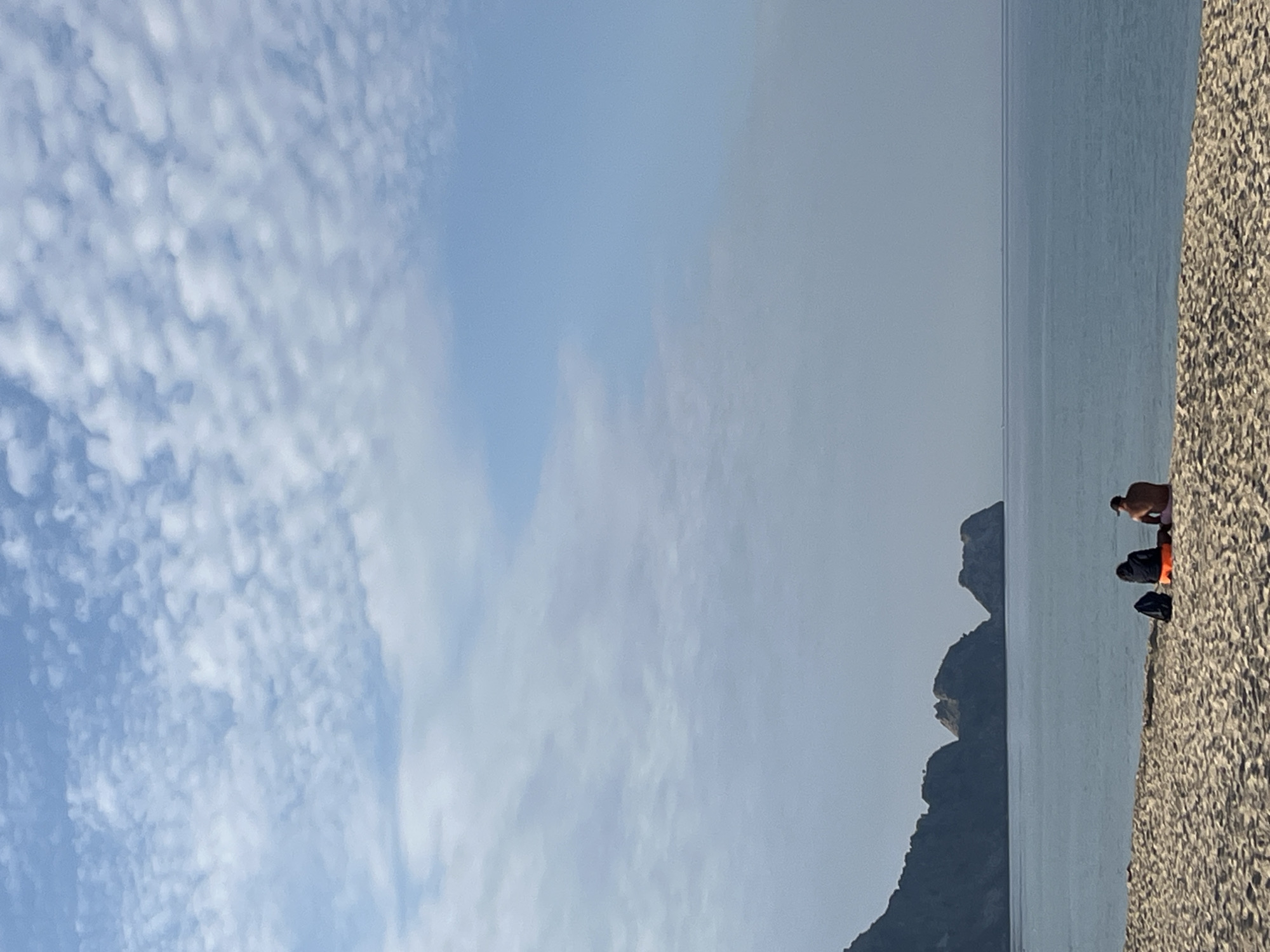 A horizontally oriented outdoor photograph depicts a coastal scene. A cloudy sky occupies the left portion of the frame. To the right, water and a rocky coastline are visible. The horizon line is approximately one-third of the way down from the top. In the lower right corner, two people are present on the beach. One person appears to be standing, while the other is seated with an orange object beside them. Additionally, there is a dark-colored backpack or bag on the beach. In the background, the silhouetted form of a land formation extends into the water, possibly a cape or peninsula. The geographical location is Alanya, Turkey.