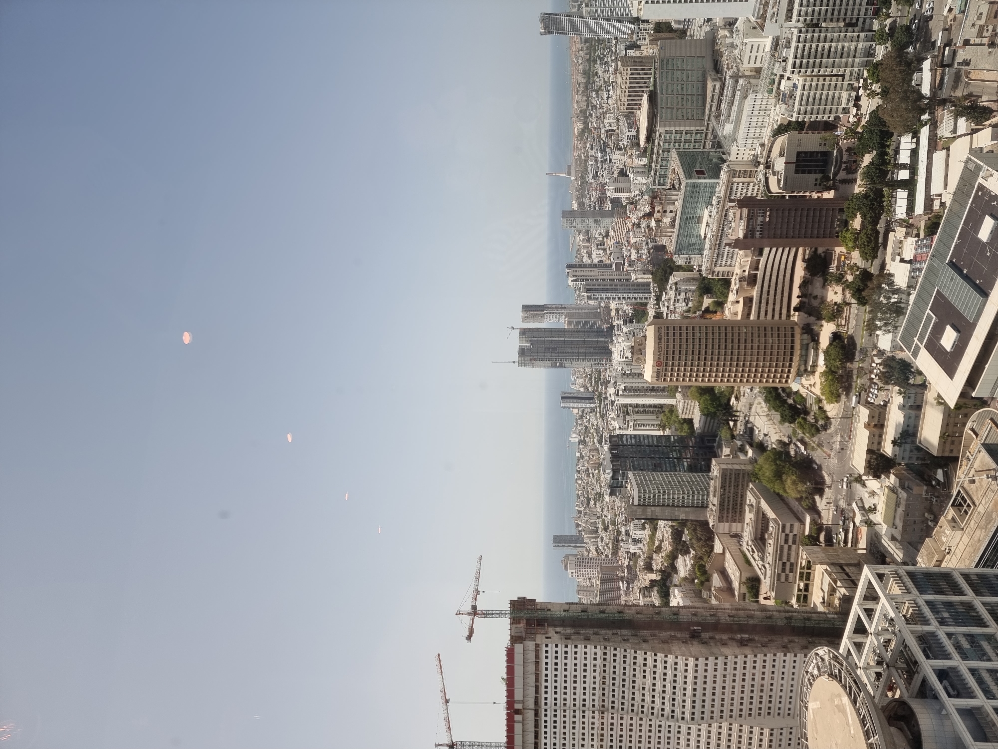 An aerial view of Tel Aviv, Israel, featuring numerous buildings, skyscrapers, and construction sites. The scene includes a clear sky with a few barely visible light red spots in the sky, the Mediterranean Sea on the horizon, and various structures in different stages of construction, with cranes visible in the foreground. The city is densely populated with buildings and roads.