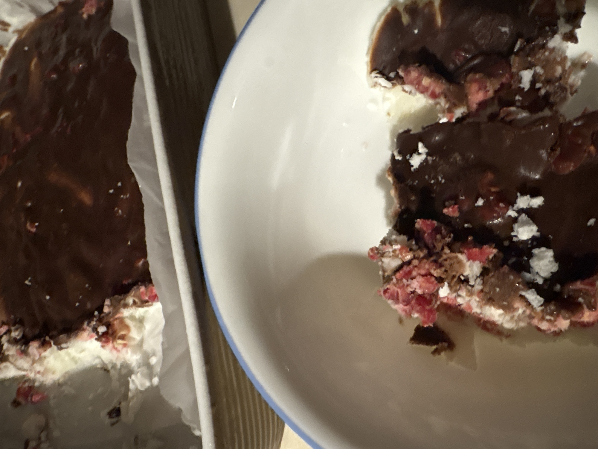 The image shows a dessert, likely a layered bar or square, partially in a white baking pan lined with parchment paper and partially served on a white plate with a blue rim. The dessert appears to have a dark chocolate topping, a middle layer with red and white specks, and a white bottom layer. The plate contains two pieces of the dessert. The scene does not contain enough information to confidently assert its location within Mexico City or if it is even located in Mexico City. There is no information about people, actions, or interactions.