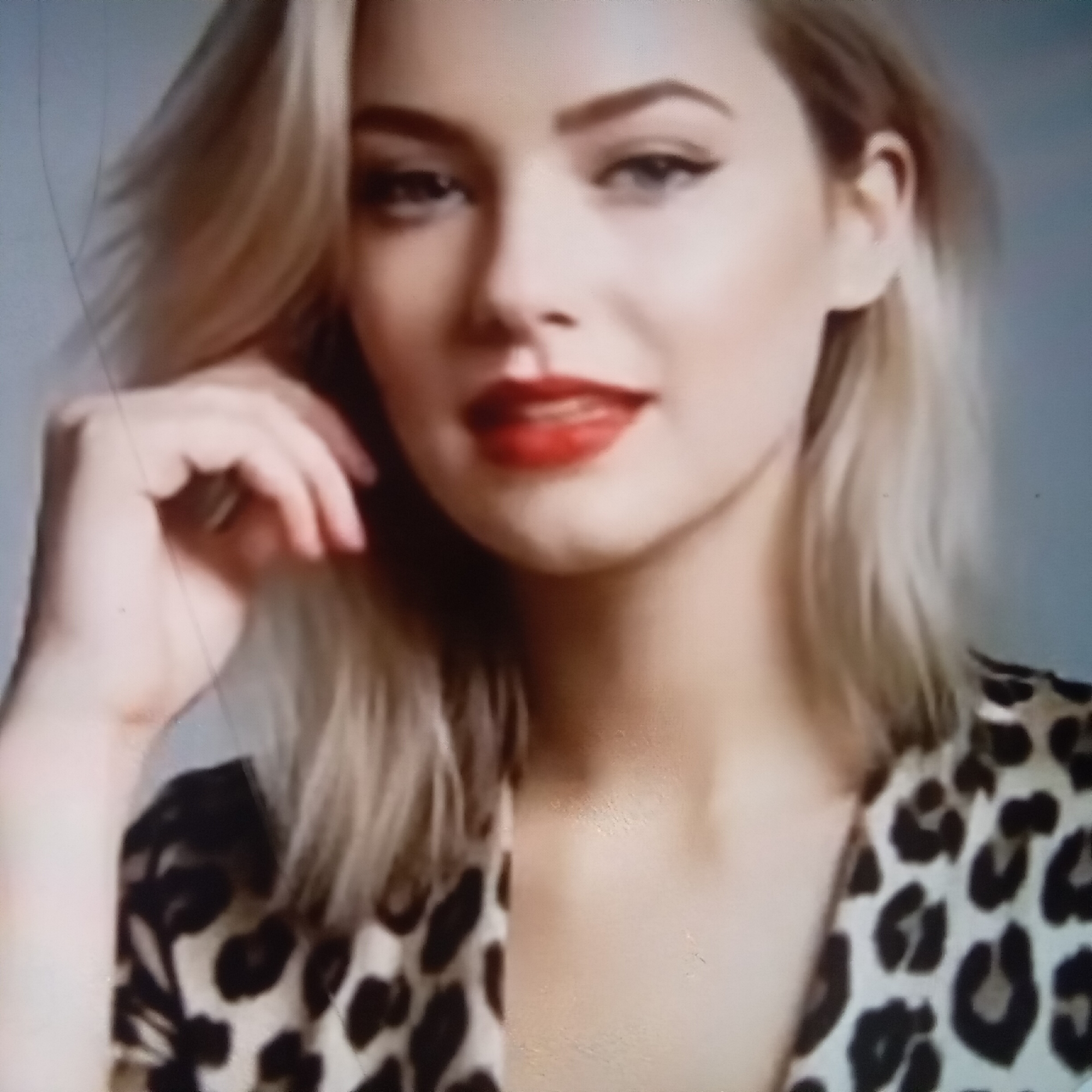 The image shows a close-up portrait of a young woman with blonde hair and fair skin. She is wearing a leopard-print top. Her eyes are made up with eyeliner, and she has red lipstick on. Her left hand is raised to her face, and she appears to be touching her cheek. The background is a muted blue-grey. There is no indication of the city of Almaty, Kazakhstan, or any other specific location within the image.