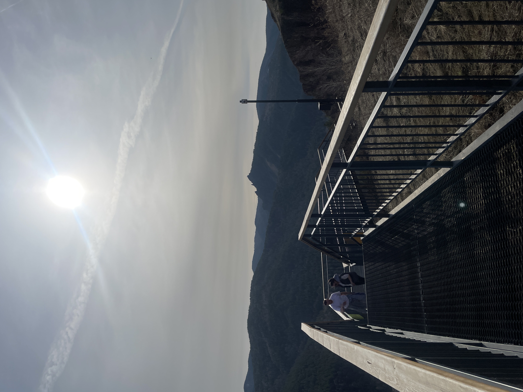 Two individuals are present on a metal and wood platform, overlooking a mountainous landscape. The platform is enclosed by railings. Above, the sky is bright with the sun and contrails. The location is Krasnoyarsk, Russia.