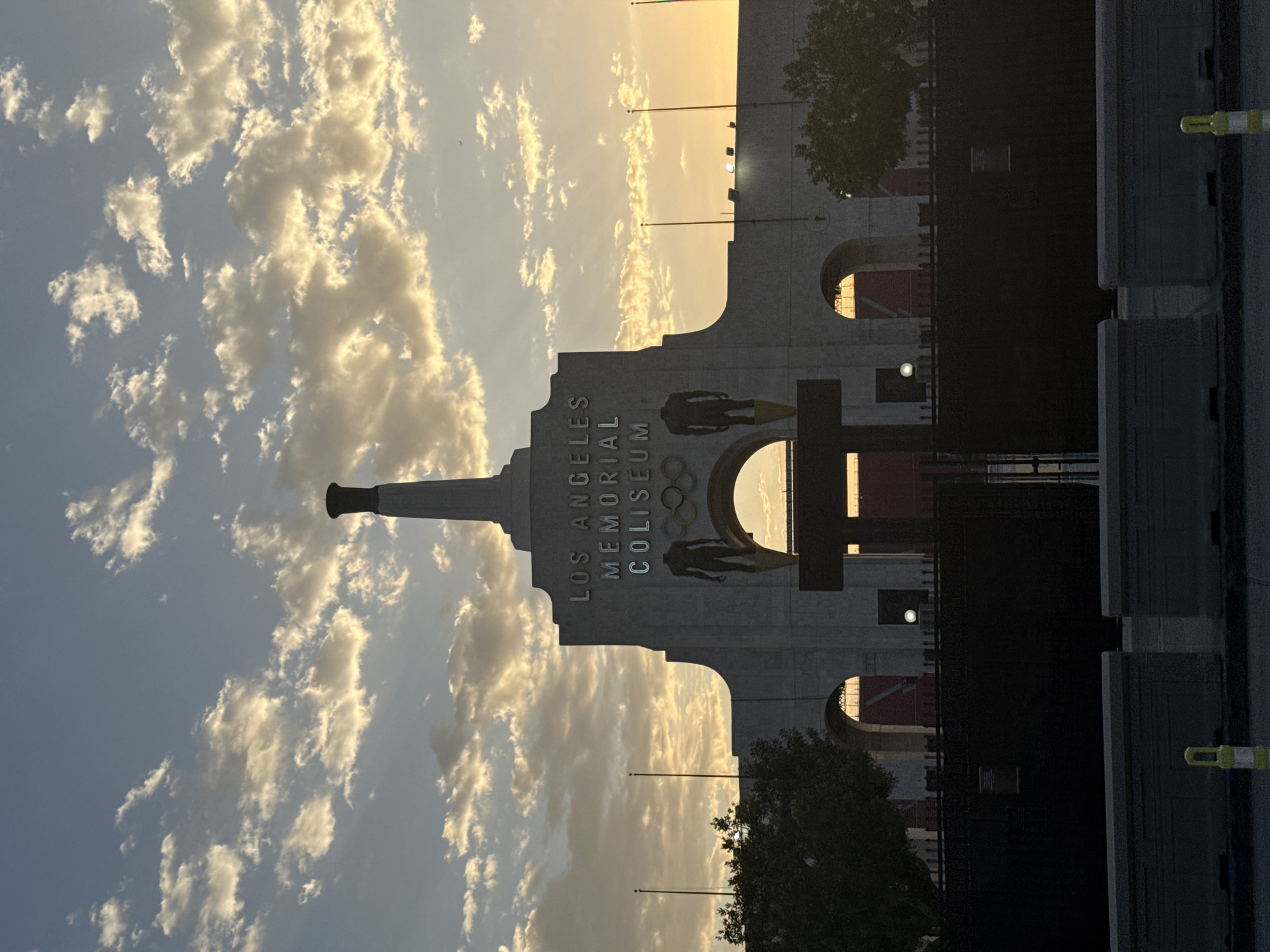 The image shows the Los Angeles Memorial Coliseum at sunset. The sky is filled with clouds, some illuminated by the setting sun, creating a dramatic effect. The iconic Coliseum structure is visible, with the words "LOS ANGELES MEMORIAL COLISEUM" emblazoned on its facade. Two statues of athletes stand prominently in front of the building, flanking an archway. Olympic rings are displayed above the arch. The foreground is dominated by a dark fence and concrete barriers, with a yellow and white traffic cone visible in the lower right corner. Trees are present on either side of the structure, and flagpoles stand tall against the sky. The overall scene captures the grandeur of the Coliseum in a twilight setting.
