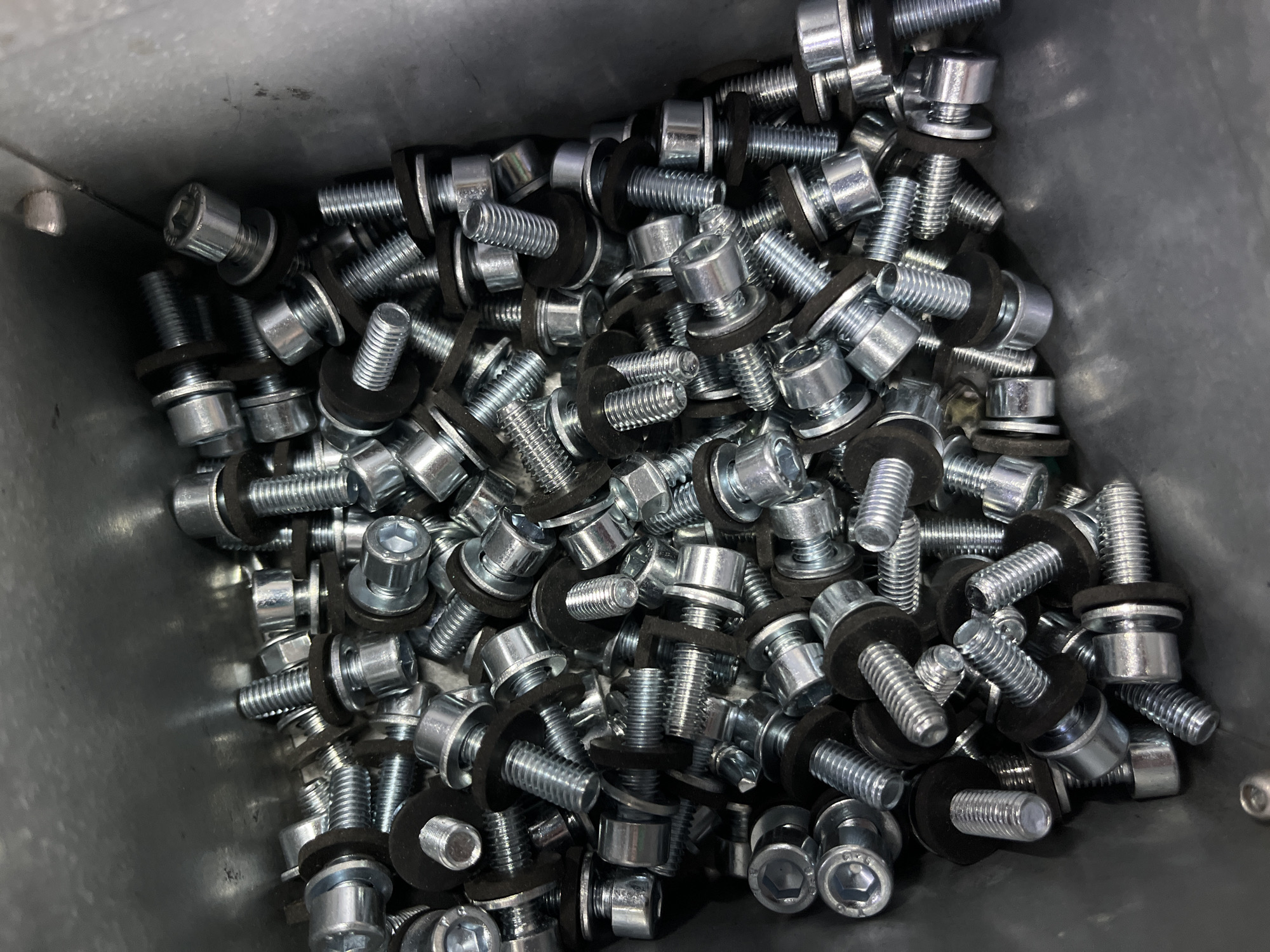 The image shows a close-up, overhead view of a large pile of silver metal bolts with hexagonal heads and accompanying dark washers, contained within a metal bin. The bolts are of various sizes and are densely packed, obscuring most of the bin's interior. The lighting appears to be artificial, casting shadows and highlighting the metallic sheen of the hardware. The scene is devoid of any human presence or activity, and the environment is industrial or storage-related, with no discernible Kyiv, Ukraine context.