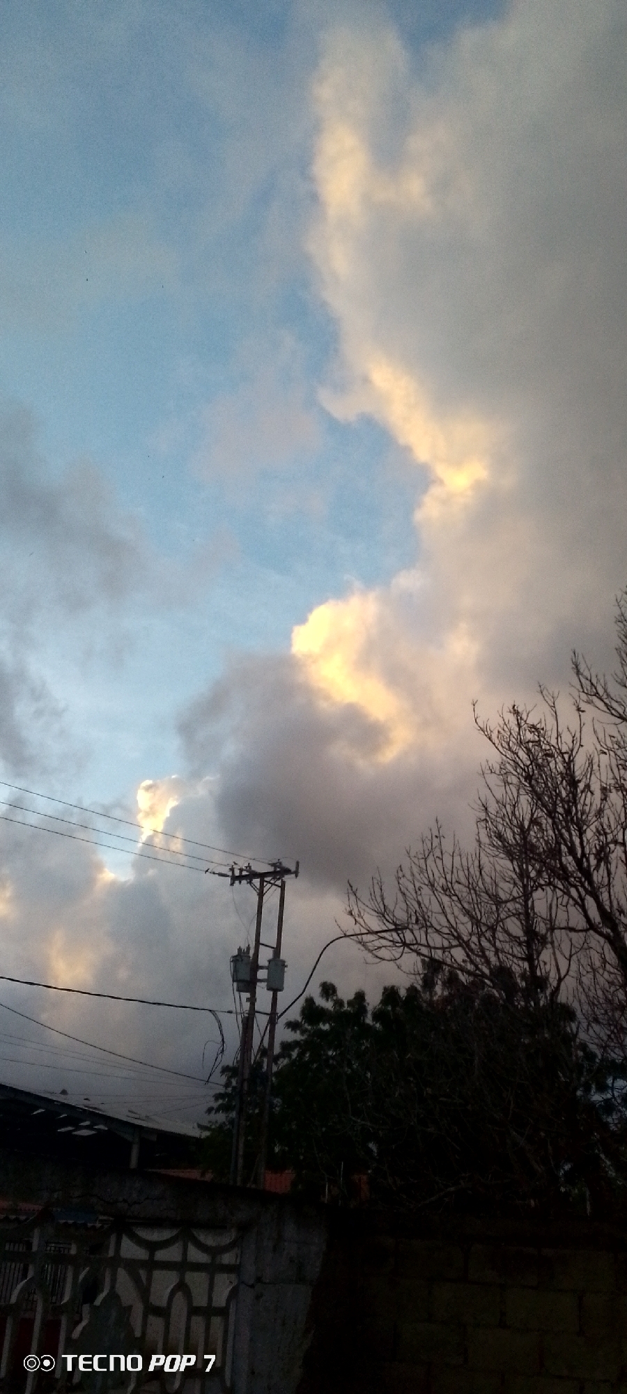 The image depicts a cloudy sky with patches of sunlight filtering through, casting warm hues on the clouds. In the foreground, a utility pole with electrical equipment is visible, along with power lines stretching across the frame. Bare tree branches, silhouetted against the sky, are present on the right side. Below the sky and power lines, a low-lying structure with a dark roof is visible. The bottom portion of the image shows a wall with an ornate pattern and the text "TECNO POP 7" in the lower left corner. The overall scene suggests an outdoor environment in a residential or urban area, possibly during a sunset or sunrise, in Altagracia, Venezuela.