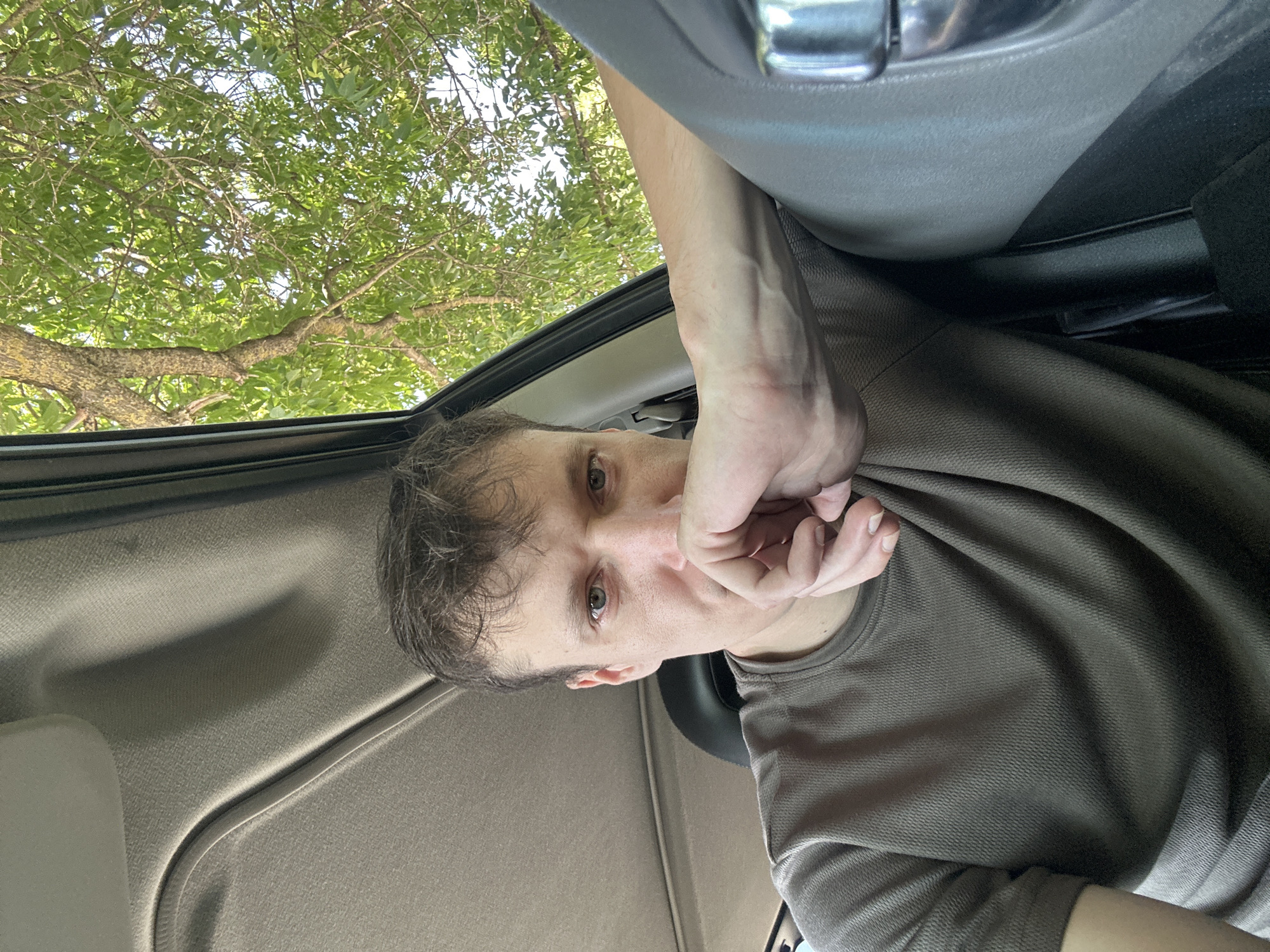 A man is captured in a close-up, selfie-style shot inside a vehicle, likely on a sunny day in Makiv, Ukraine. He is seated in the driver's seat, with his left arm resting on the steering wheel. His gaze is directed towards the camera, and his right hand is raised to his face, with his knuckles against his mouth, creating a thoughtful or contemplative expression. The interior of the car is visible, including the car's headliner and a portion of the dashboard. Through the open car window, lush green tree branches and leaves are visible against a bright sky, suggesting an outdoor, possibly rural or suburban, setting. The man is wearing a plain, short-sleeved t-shirt. The lighting indicates it is daytime.
