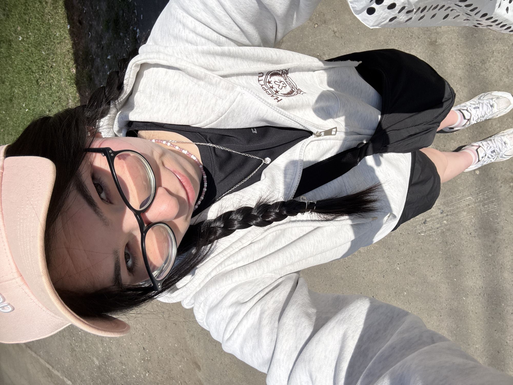 A woman wearing a light pink baseball cap and glasses is taking a selfie in an outdoor setting. She is wearing a grey zip-up hoodie over a black shirt, black shorts, and white sneakers with pink striped socks. She has a braid in her dark hair and is wearing necklaces. The background shows a concrete surface and grass. The location is not identifiable as Izhevsk, Russia based on the visual information.