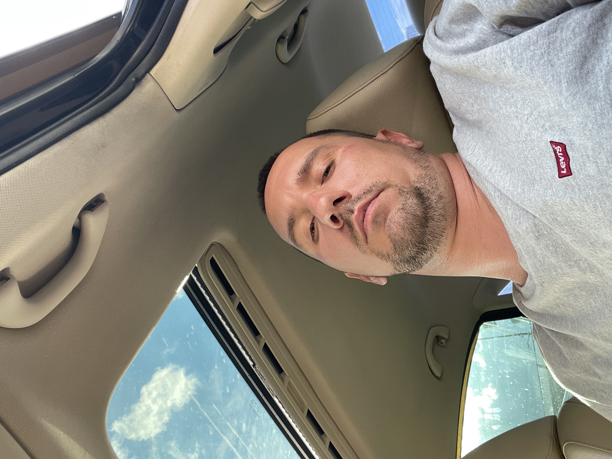 A selfie of a man taken from inside a car, looking up towards the sunroof. He is wearing a grey sweatshirt with a red Levi's logo on the left chest. The sunroof is open, revealing a blue sky with white clouds. The interior of the car is visible, including the beige headliner and a portion of a beige seat. The man has short brown hair, stubble, and appears to be in his 30s or 40s. There are no identifiable landmarks indicating a specific location in Kyiv, Ukraine.