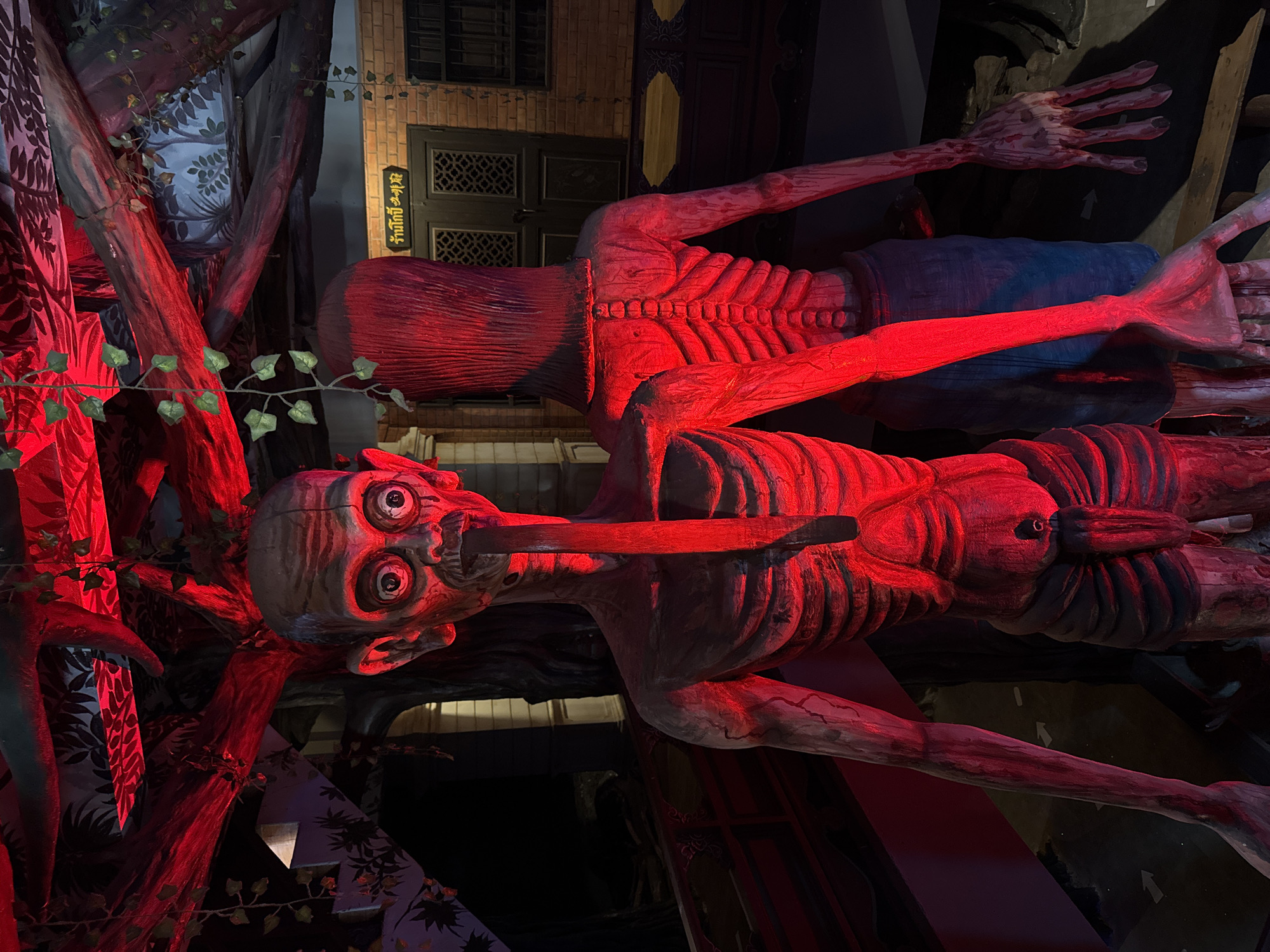 The image shows a distorted, red-lit scene, likely a display or installation, located in Bang Pu Subdistrict Municipality, Thailand. Two skeletal figures are prominently featured. One figure has an elongated face with exaggerated eyes and a stick protruding from its mouth. Its skeletal rib cage is visible. The other figure is positioned behind and above, also displaying a prominent rib cage. A sign written in Thai is visible in the background above the upper skeleton. The surrounding environment includes stylized trees and drapery, with red lighting bathing the entire scene. The scene appears to be theatrical or part of a themed environment.