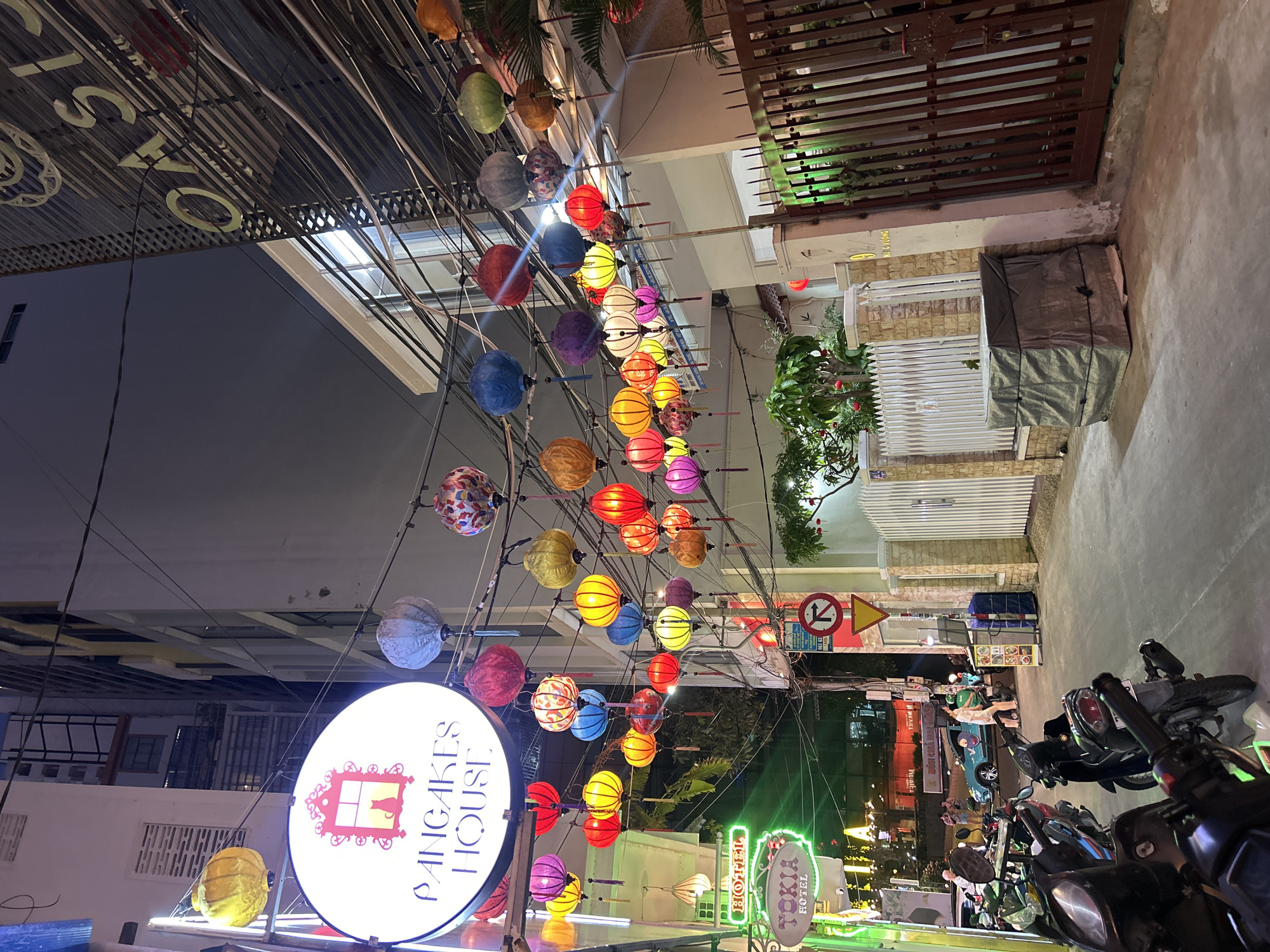 The image shows a street scene in Nha Trang, Vietnam, at night. A row of colorful lanterns hangs across the street, illuminated. On the left, there's a sign for "Pancakes House" with an illustration. On the right, several motorbikes are parked along the sidewalk. Pedestrians are present further down the street. There is a sign of "no entry for vehicles" in view. Buildings line both sides of the street, and various signs and lights indicate commercial establishments.