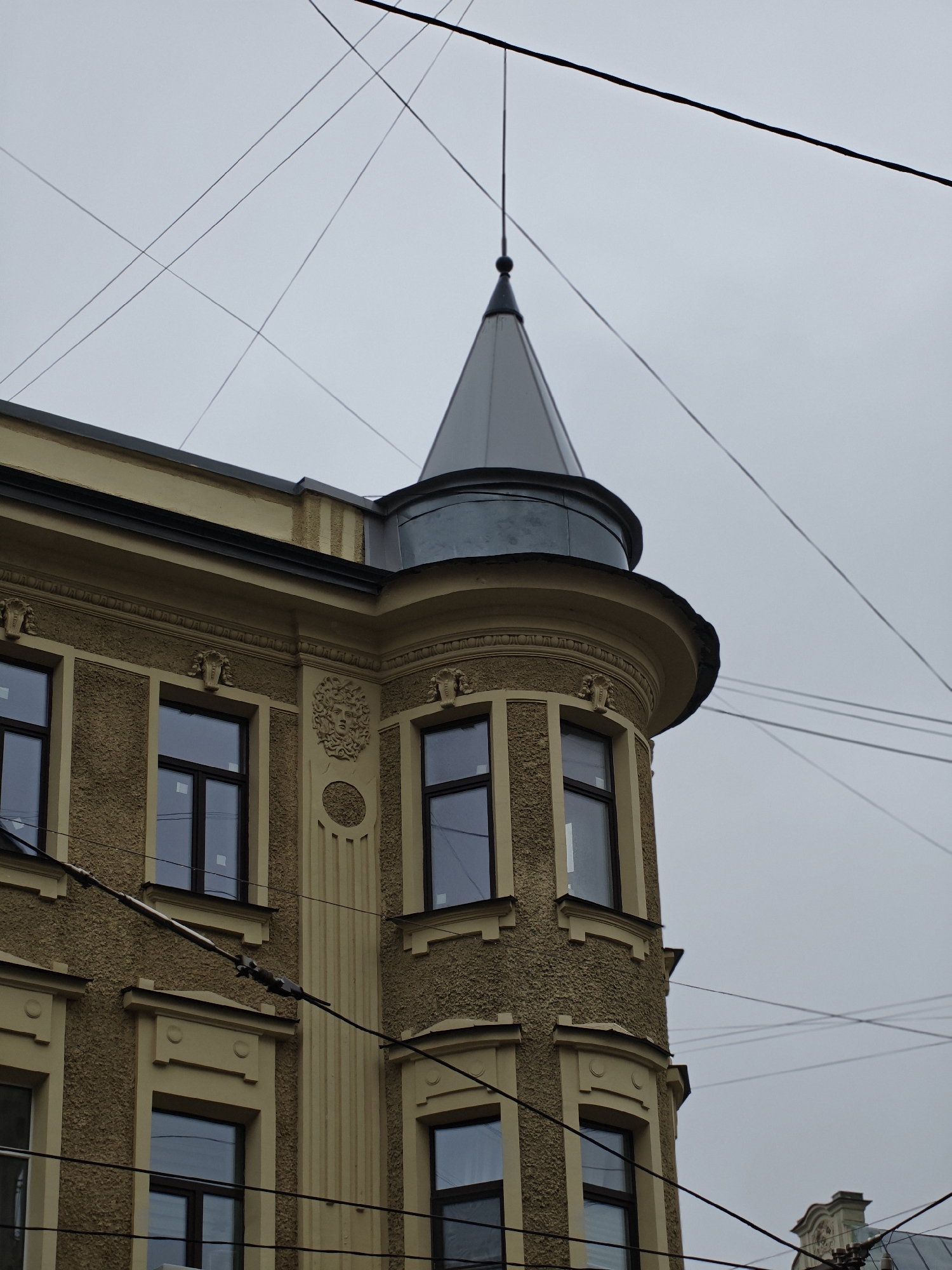 A low-angle, close-up shot features the upper corner of a yellow, stucco-clad building in an urban environment. The building has a distinct turret with a conical, dark gray roof, topped with a thin, dark spire. Multiple windows with dark frames are visible on the building's facade, some appearing to have white coverings or films on the glass. The building exhibits architectural ornamentation, including relief sculptures above some windows and a decorative frieze. The scene is dominated by a complex network of dark, thin wires crisscrossing against a muted, overcast sky. The wires appear to be overhead electrical or communication lines, and some are attached to the building via insulators. The overall impression is one of an old European city with a sense of urban density and perhaps a slightly melancholic or muted atmosphere due to the cloudy sky.