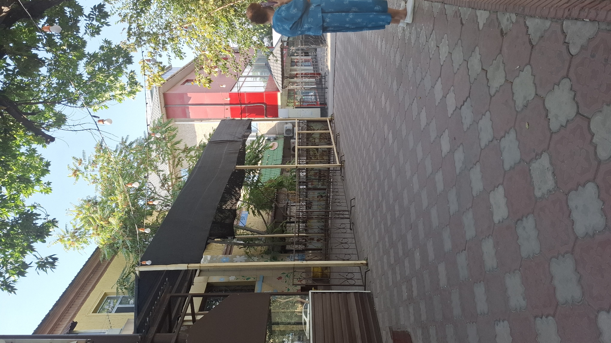 A woman in a blue dress stands on a patterned brick walkway, looking at her phone. She is facing a building with a red facade and a black awning. The building is surrounded by trees and string lights are visible overhead. The scene appears to be an outdoor seating area for a restaurant or cafe in Shymkent, Kazakhstan.