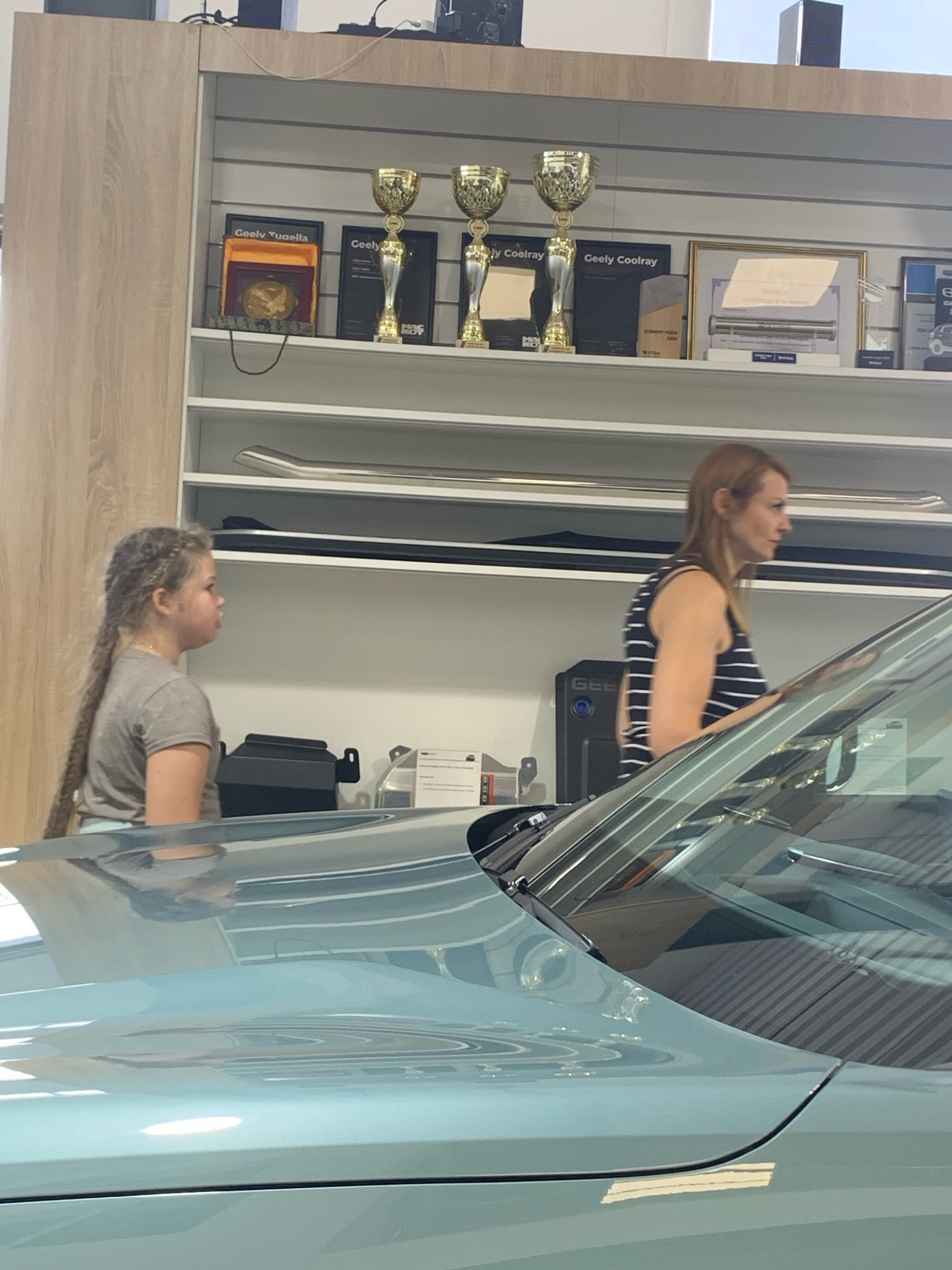 The image shows an indoor scene, likely a car dealership, with a light-colored wooden cabinet on the left and white shelves on the right. On the shelves, there are awards and framed documents. In the foreground, a light blue car is partially visible, with its hood and windshield taking up a significant portion of the frame. Two people are present: a young girl with long braided hair is standing on the left, facing the right. A woman with shoulder-length reddish-brown hair, wearing a black and white striped sleeveless top, is standing on the right, also facing right. She appears to be looking towards the car. The environment suggests a commercial setting. The location is identified as Чыжэвіцкі сельскі Савет, Belarus, which is a rural administrative region in Belarus.