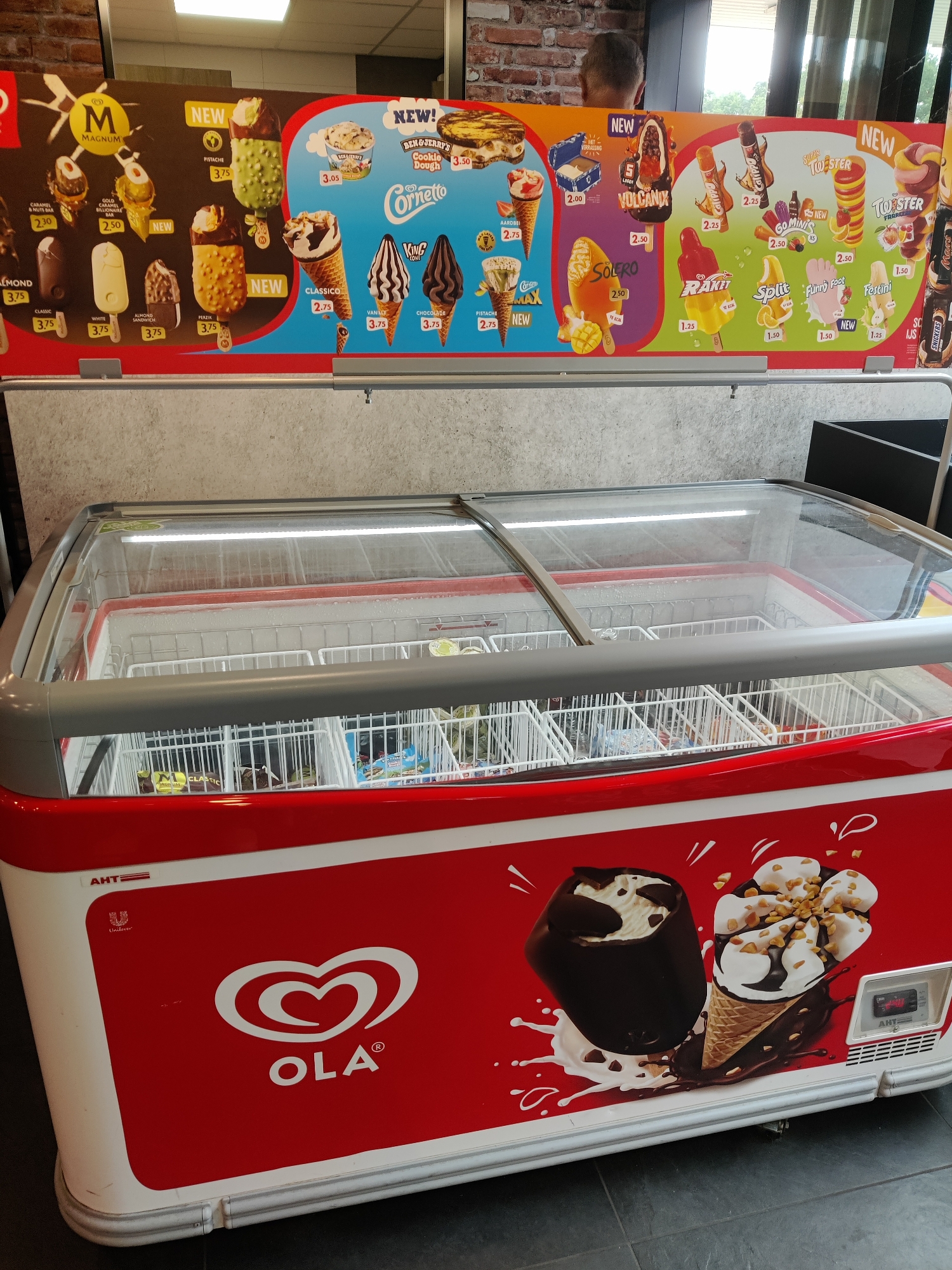 The image shows an indoor scene at an unknown retail establishment in Eijsden, Netherlands, featuring a red and white chest freezer stocked with various ice cream products. The freezer has a glass lid that is partially frosted, indicating its cooling function. Above the freezer, a colorful advertisement poster is displayed, showcasing different ice cream brands and flavors with prices. The poster includes recognizable brands like Magnum, Cornetto, Ben & Jerry's, and Ola. The environment appears to be clean and well-lit, with tiled flooring visible. The overall impression is of a point-of-sale display in a shop or supermarket.