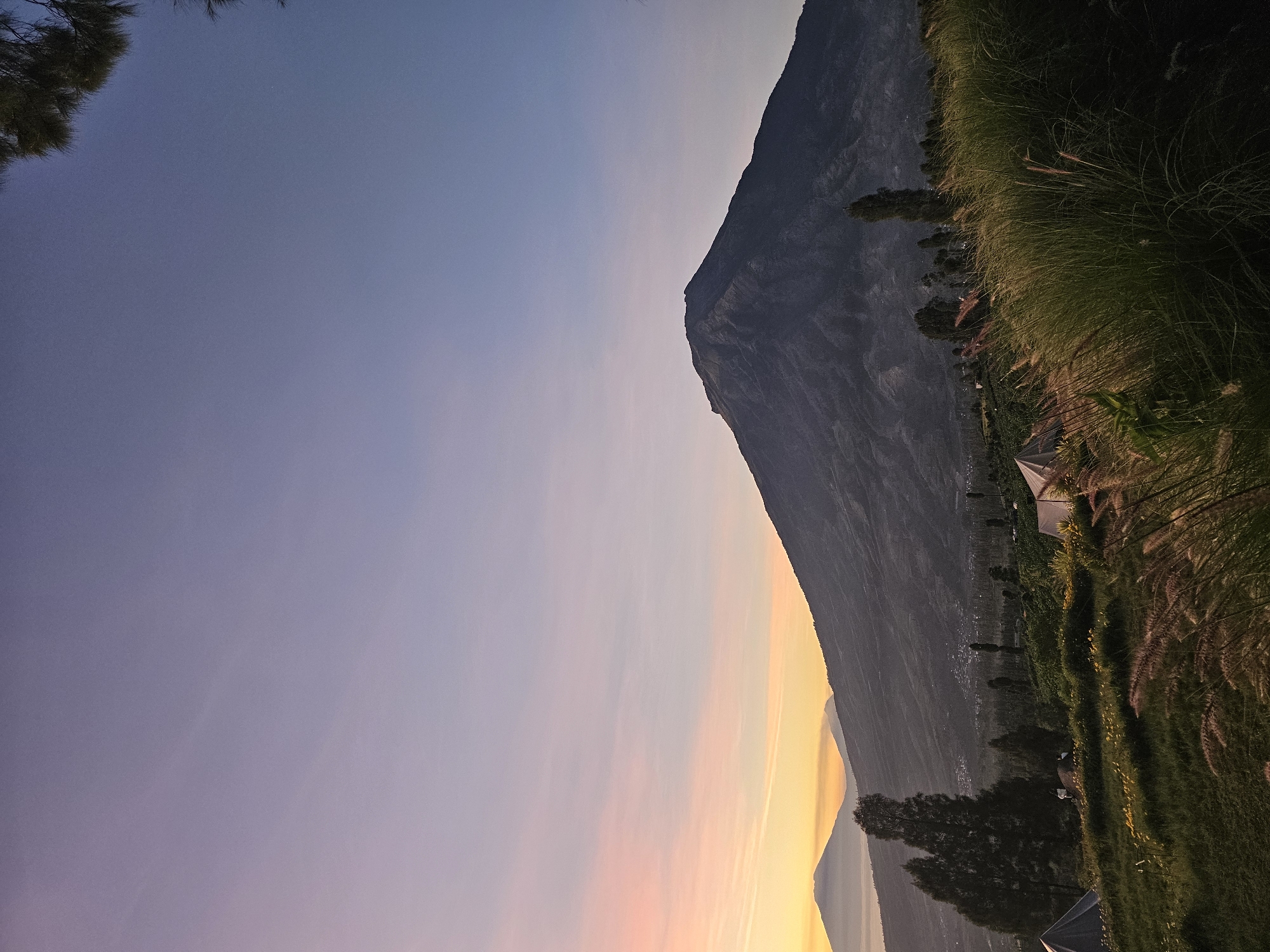 In Tlahab, Indonesia, a mountain landscape is captured during twilight. A large, dark mountain dominates the right side of the frame, its slopes textured with shadow and light. In the distance, two smaller, hazy mountains are visible against a pastel sky transitioning from soft orange and pink near the horizon to a pale lavender and blue above. The foreground features a grassy slope dotted with wildflowers, with several tents set up for camping. Tall, wispy grasses in the immediate foreground partially obscure the view, adding a sense of depth and texture to the scene. Scattered trees and vegetation populate the middle ground, leading down to a distant, lightly populated valley. The overall atmosphere is serene and tranquil, indicative of a peaceful natural setting at dawn or dusk.