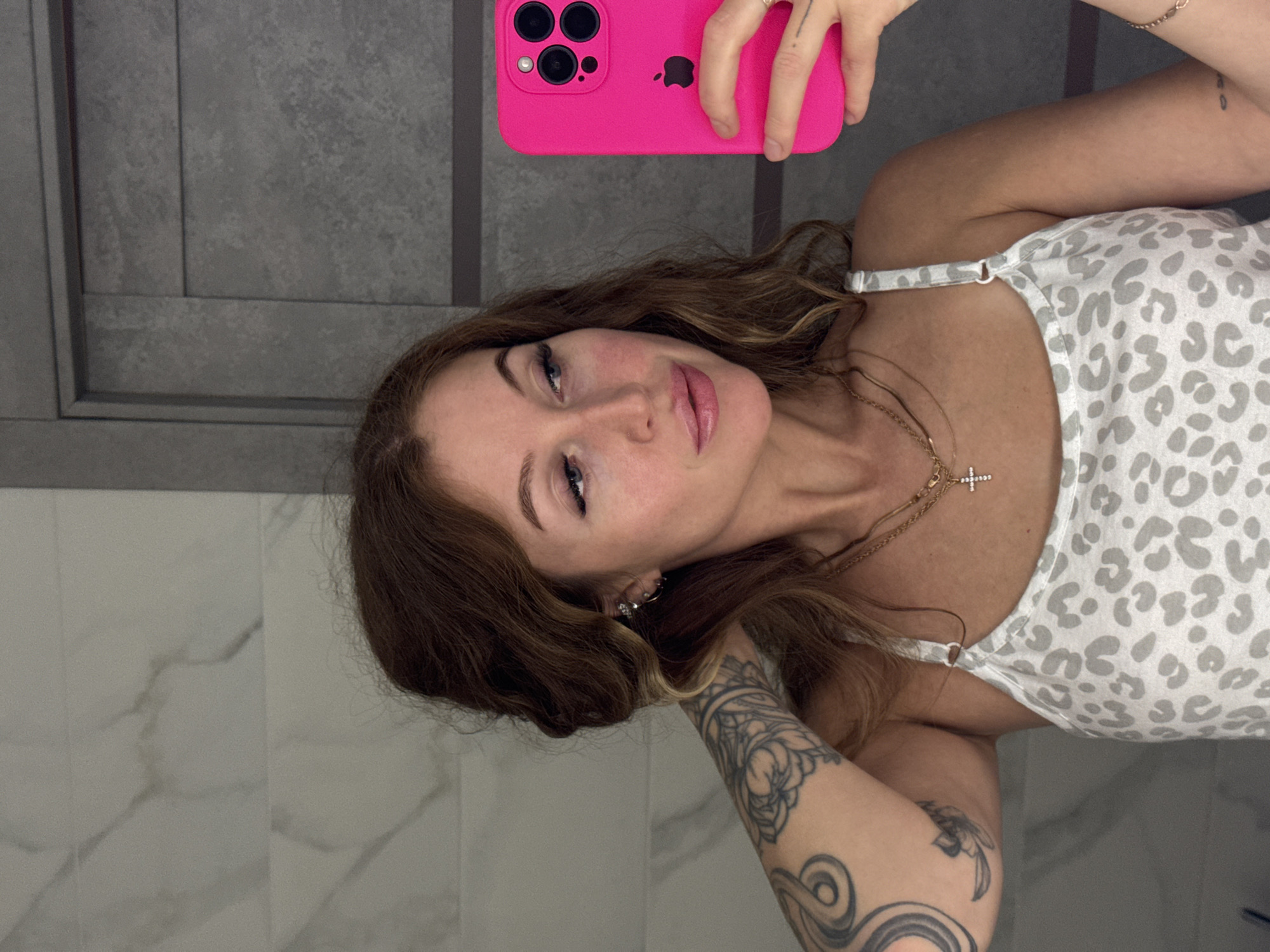 A woman with shoulder-length brown hair is taking a selfie in what appears to be a bathroom. She is holding a bright pink iPhone in her right hand. She's wearing a white and grey leopard print tank top. She has multiple tattoos on her left arm and is wearing a necklace with a cross pendant. The bathroom has grey paneling and white marble-look tiles. There is no evidence indicating the image was taken in Chaikovsky, Russia.