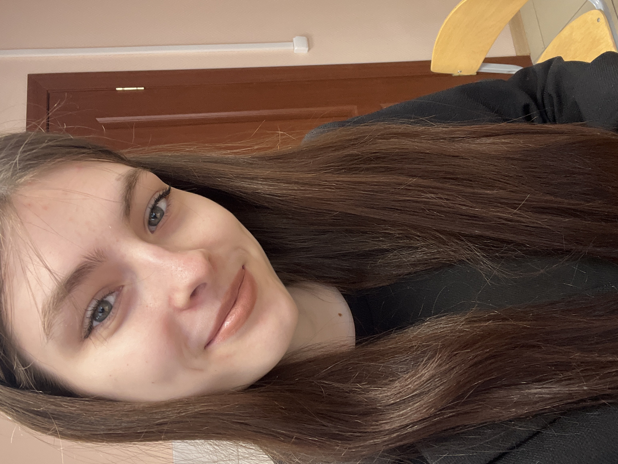 A close-up, indoor photograph features a young woman with long, brown hair. Her face is turned slightly towards the viewer, displaying her features: fair skin, light-colored eyes, and a soft, neutral lip color. She appears to be wearing a black garment.
The background includes a brown door or panel and part of a light-colored wooden chair. The location, Saint Petersburg, Russia, cannot be determined from the visual information provided. The scene lacks specific details that could confirm the location with certainty.