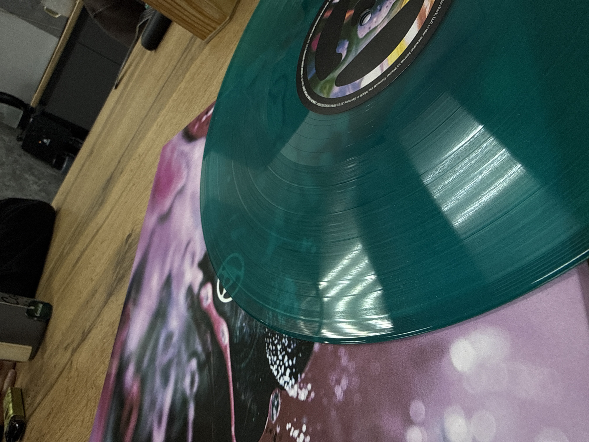 A close-up shot depicts a turquoise vinyl record resting on a record cover. The record cover features a blurry image with purple, pink, and black colors and bokeh light effect. The edges of the record cover are visible at the bottom and left side. A portion of the record label is seen on the top right, with text visible along the edge of the label. The vinyl record is shiny and reflective. The surface underneath is likely a wooden surface. The location of Chaikovsky, Russia is not evident from the image.