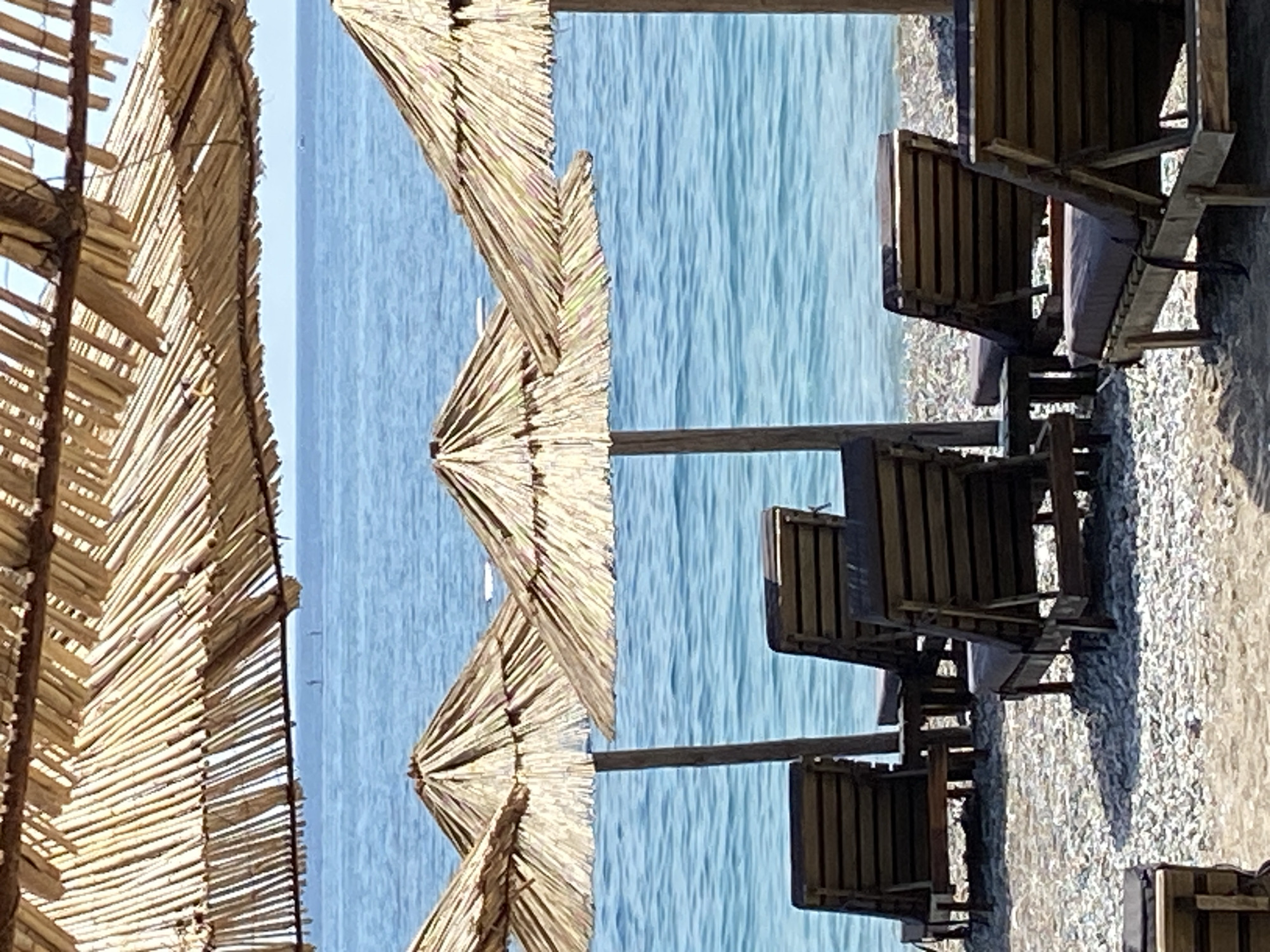 This image depicts a beach scene at Prijevor, Montenegro. The foreground is dominated by thatched beach umbrellas, made of natural materials like straw or palm leaves, with wooden frames. These umbrellas are arranged in rows, suggesting a public beach area. Below the umbrellas, several wooden lounge chairs are visible, some with cushions. The chairs are made of weathered wood, indicating outdoor use.
Beyond the beach furniture, a body of calm, blue water stretches to the horizon. The water appears to be the Adriatic Sea, given the location context. The surface of the water is smooth with slight ripples, suggesting a calm day. The sky is a pale blue, indicating clear weather.
The image appears to have been taken during daylight hours, likely in the morning or afternoon, as there are strong shadows cast by the umbrellas and chairs. The overall mood of the image is one of relaxation and tranquility, characteristic of a beach vacation. There is no visible text in the image. The scene is set on a pebble beach, indicated by the texture of the ground beneath the chairs. The composition is such that the sea forms a significant backdrop to the beach amenities.