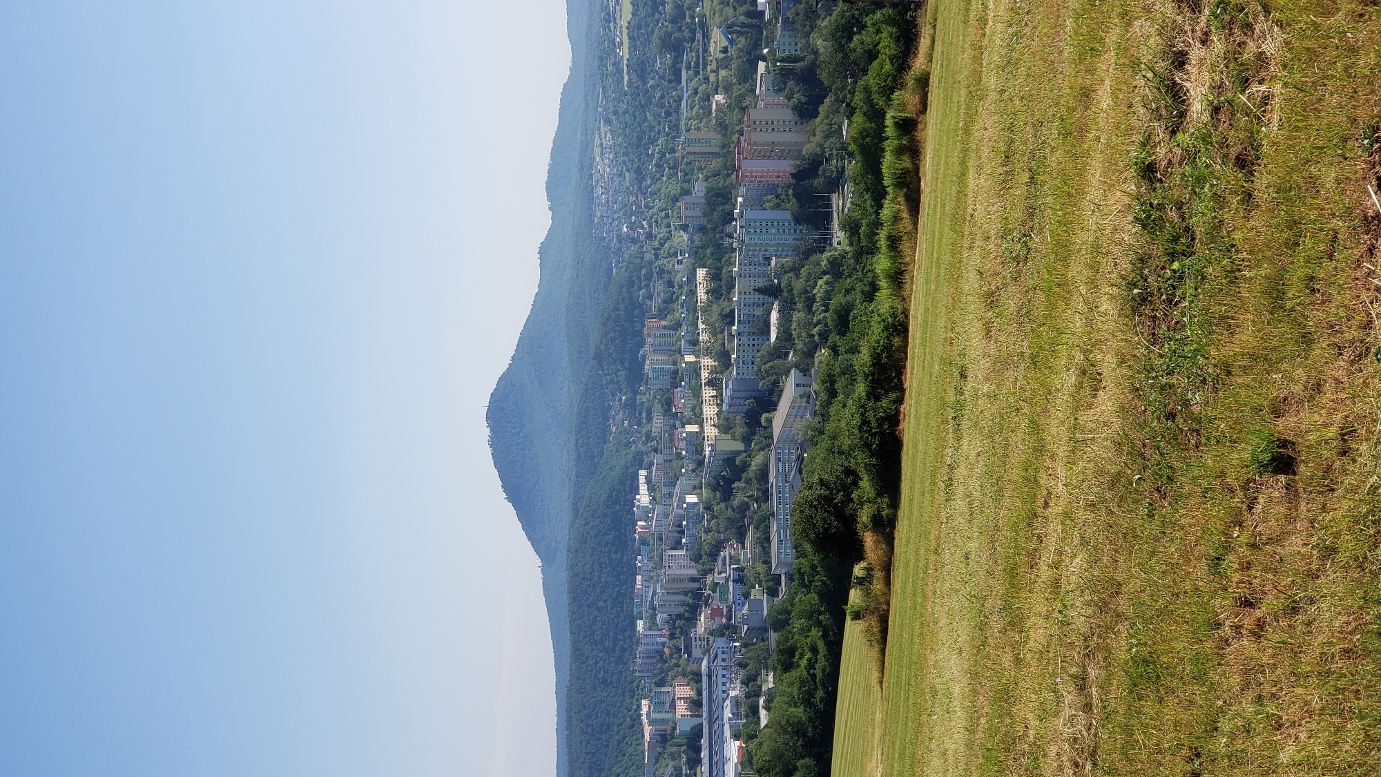 The image displays a panoramic view of the city of Prešov, Slovakia, under a clear blue sky. In the foreground, a gently sloping field covered in dry and green grass occupies the lower portion of the frame. The field appears to be recently mown, with distinct lines visible in the grass.
Beyond the field, a densely populated urban area stretches across the middle ground. Numerous apartment buildings, primarily characterized by their concrete construction and multi-story design, dominate the cityscape. These buildings exhibit a variety of colors, including shades of white, yellow, pink, and grey. Scattered among the residential structures are trees and patches of greenery, indicating a balance between urban development and natural elements. Industrial or commercial buildings with flat roofs and solar panels are also visible on the left side of the city.
In the background, a prominent conical hill, known as Šariš Castle Hill, rises majestically. The hill is covered in lush green vegetation, and a telecommunications tower or observation post is discernible at its summit. The surrounding landscape is characterized by rolling hills and dense forests, creating a picturesque backdrop for the city. The overall impression is one of a well-established urban center nestled within a verdant natural environment. No people or specific actions are discernible in the image.