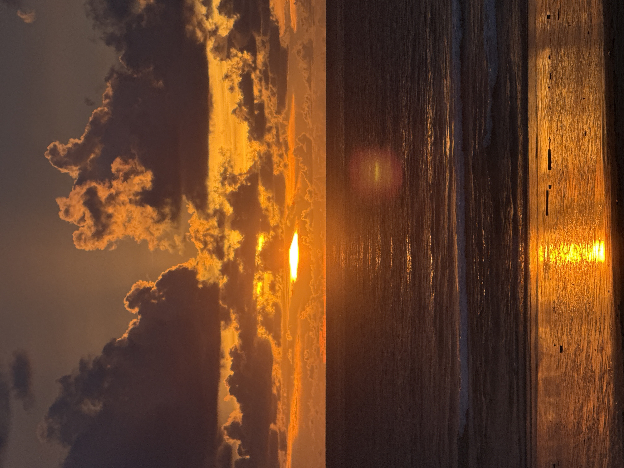 An extreme wide shot captures a dramatic sunset over the ocean. The sun, a bright orange disc, is partially obscured by a thick band of clouds, casting a warm, golden glow across the scene. The clouds are a mix of dark gray and vibrant orange, creating a striking contrast. The ocean is a deep blue-gray, with gentle waves lapping at the shore. The wet sand on the beach reflects the sunset, creating a shimmering expanse of orange and gold. The image is taken from a slightly elevated perspective, looking out over the water and towards the horizon. The scene is devoid of any human presence.