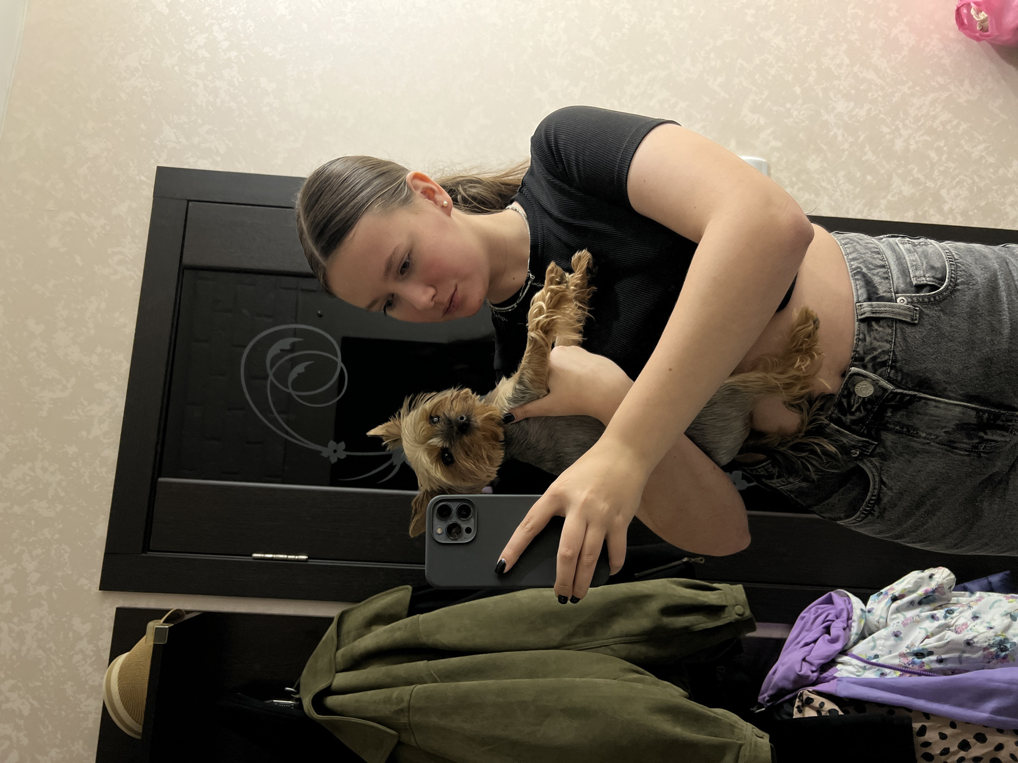 A young woman is holding a small dog in her arms and taking a selfie in front of a mirror. The woman is wearing a black crop top and gray jeans. The dog is a Yorkshire Terrier. The mirror is reflecting a wardrobe, on which there is a green jacket and a pile of colorful clothing. The wall behind the mirror is wallpapered with a beige pattern. The location is Chaikovsky, Russia.