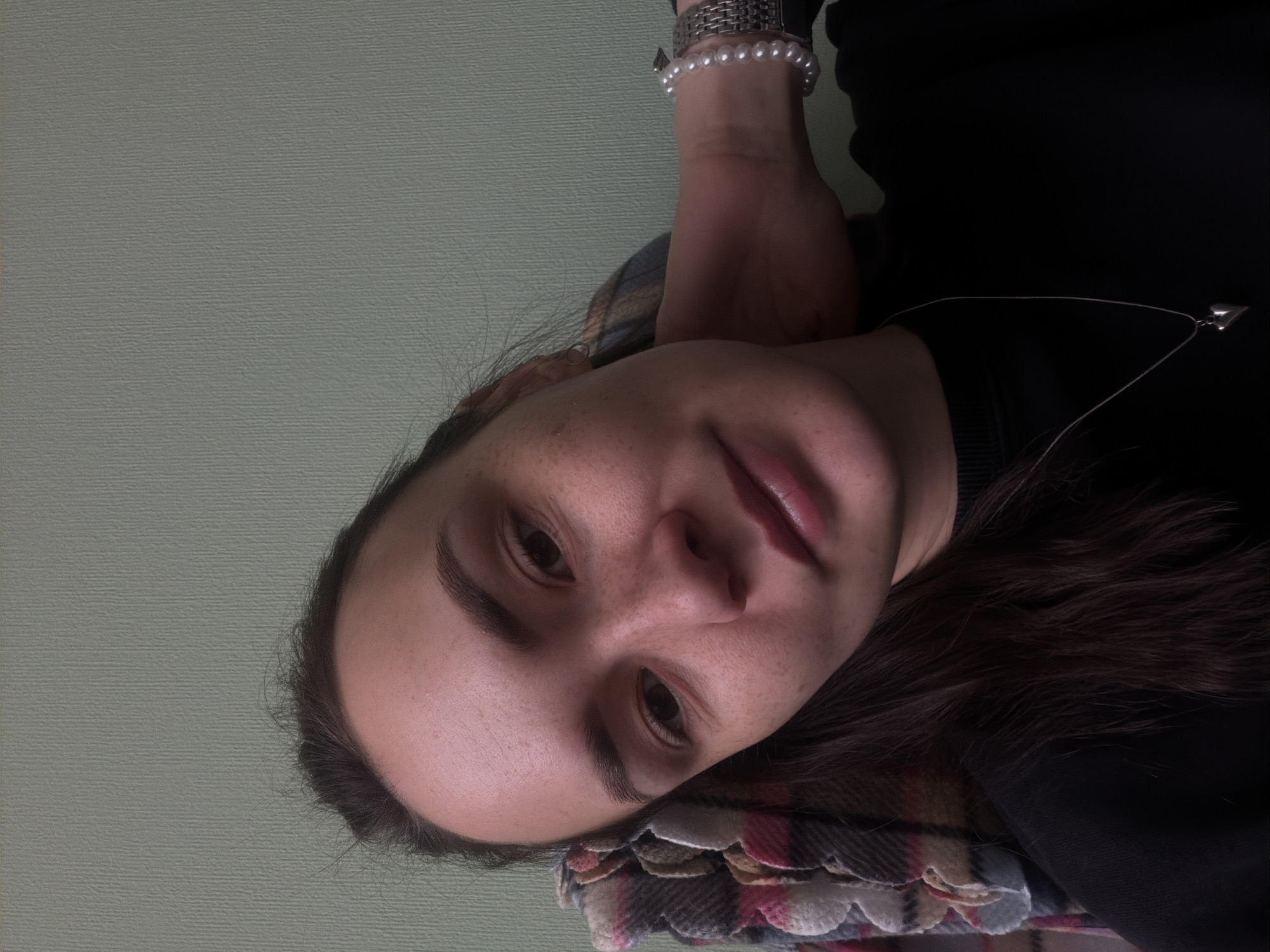 A close-up, slightly angled portrait shot depicts a fair-skinned person with dark brown hair, lying down. The person has visible freckles. They are wearing a black top and a silver necklace with a heart-shaped pendant. A silver watch with pearl details is visible on their wrist. The person is lying on a plaid blanket. The background is a pale green wall. There is no verifiable information to suggest this image was taken in the city of Chaikovsky, Russia.