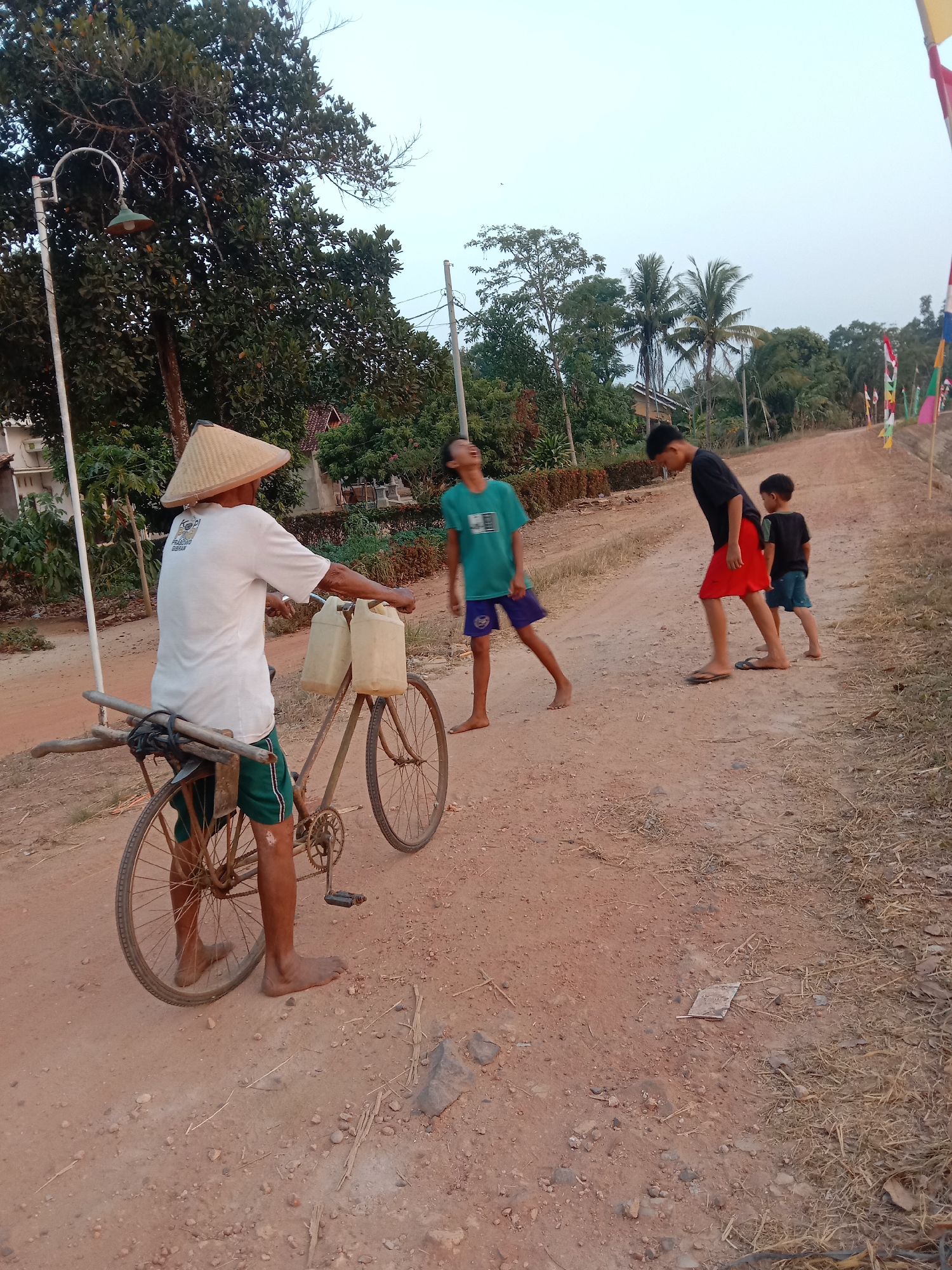 An outdoor, eye-level, medium shot captures a rural scene with four individuals and a bicycle on a dirt road. In the foreground, a man wearing a conical straw hat and a white t-shirt with green shorts rides a weathered bicycle. Two light-colored plastic jugs are attached to the bicycle's frame, and a bundle of wooden poles is secured to the rear. The man is barefoot. To the right of the man, three boys are present. One boy in a teal t-shirt and blue shorts stands looking up. Another boy in a black t-shirt and red shorts stands with his head down, and a third boy in a black t-shirt and blue shorts stands looking ahead. The environment is characterized by a reddish-brown dirt road, dry grass, and scattered stones. Lush green trees and some foliage line the sides of the road, with houses visible in the background. Flags are strung along the right side of the road in the distance. The sky is a pale, hazy blue, suggesting either early morning or late afternoon. The overall atmosphere is that of a humble, possibly agrarian, community.
