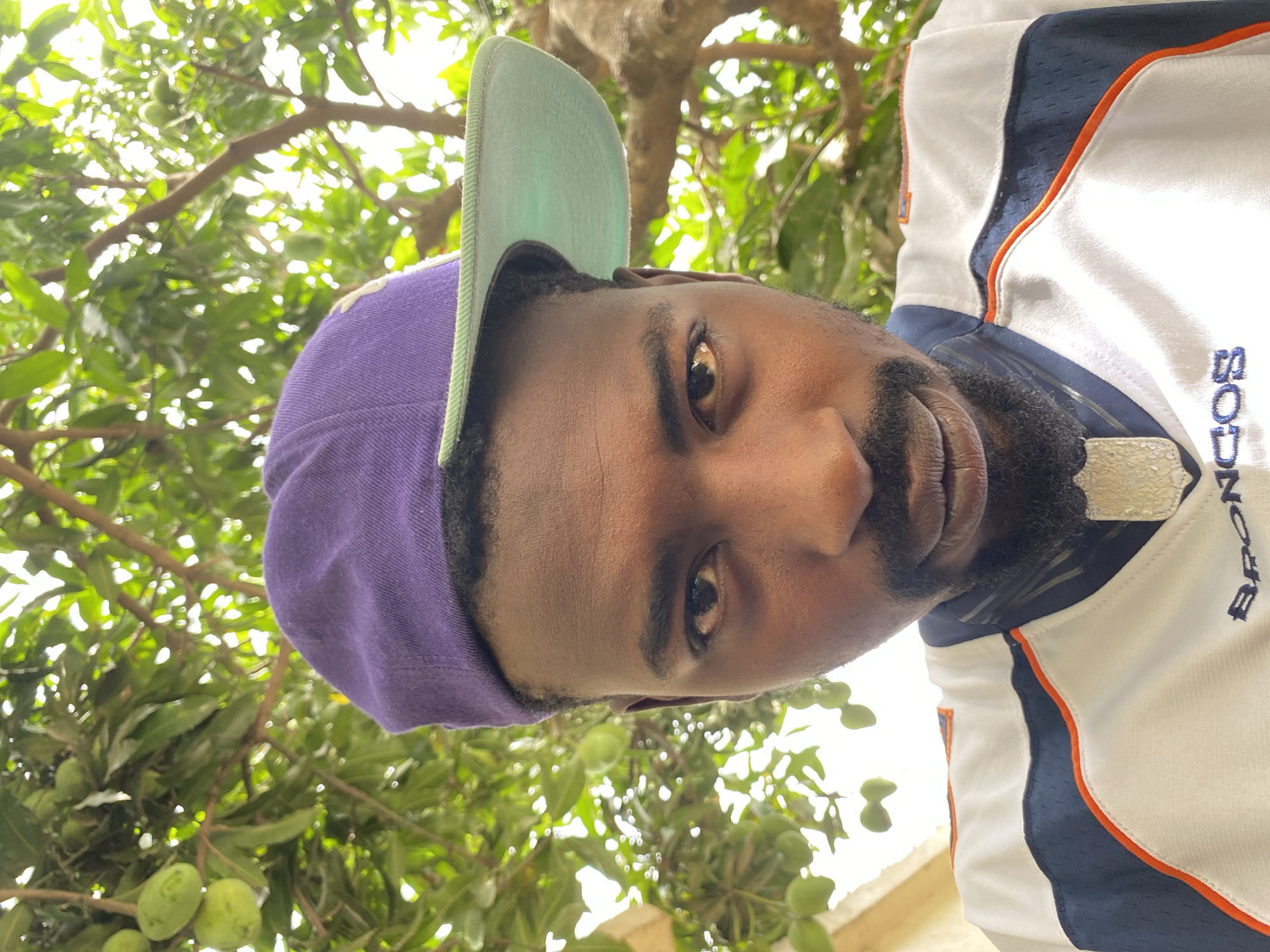 A portrait-style, upward-angled, and rotated photograph shows a Black man wearing a purple and green baseball cap and a white jersey with blue and orange trim. The jersey has the word "BRONCOS" printed vertically. The man has brown eyes and a dark beard. The background features green leaves and small green fruits hanging from branches, suggesting a tree canopy. The scene implies an outdoor setting, possibly in Barakin Akawo, Nigeria, where tropical fruit trees might be common. The photograph lacks any explicit actions or interactions; it is a still image focused on the man.
