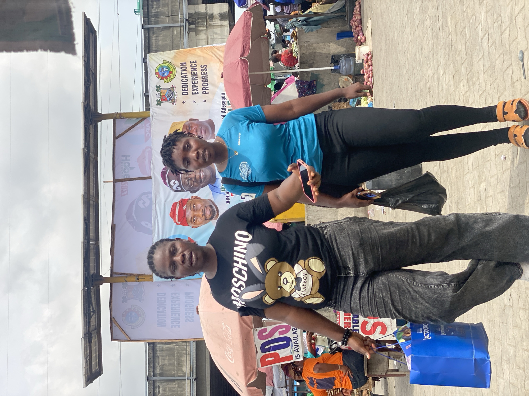 This image, presented vertically, captures an outdoor street scene in Lekki, Nigeria, characterized by market activity under an overcast sky.
In the foreground, two women are posing for the camera. The woman on the left wears a black t-shirt featuring "MOSCHINO" and a graphic of a Playboy teddy bear, paired with dark grey jeans. She holds a blue shopping bag in one hand and a mobile phone in the other. The woman on the right is dressed in a light blue t-shirt with "CRAFT" and a circular logo, black leggings, and orange sandals, also holding a mobile phone.
The background reveals a bustling market environment. Piles of red onions are displayed on the paved ground near vendor stalls, along with a large water dispenser. A red umbrella, branded with "Coca Cola," provides shade over a stall, while another brown umbrella is visible further back. Overhead utility wires stretch across the cloudy sky.
Several banners and signs contribute to the scene's context. A prominent, large banner features portraits of multiple men, including names like "Rola" and "d Adesegun," alongside campaign slogans such as "* DEDICATION * EXPERIENCE * PROGRESS" and "ETIOSA LOCAL GOVERNMENT." Another sign announces "POS IS AVAILAB," indicating the availability of point-of-sale services. The blue shopping bag held by the woman on the left also displays text including "LACTOGRO." The overall impression is that of a lively urban commercial area during the daytime.