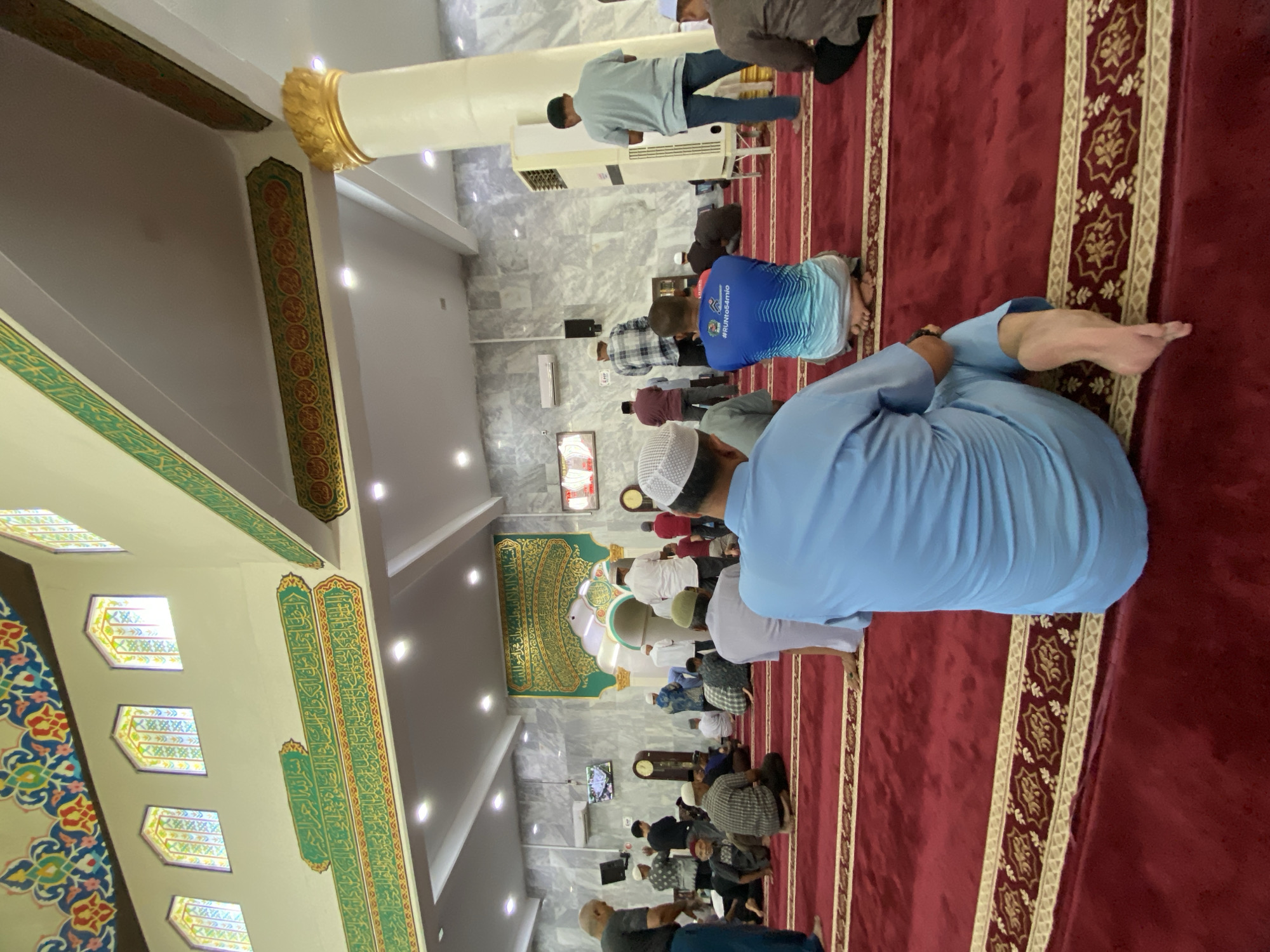 The image depicts the interior of a mosque in Lhokseumawe, Indonesia, filled with Muslim worshippers. The mosque features a red carpet with a decorative border, high ceilings adorned with intricate Arabic calligraphy and colorful geometric patterns, and ornate gold-trimmed pillars. Several men are visible, most of them wearing traditional Muslim attire such as peci caps and long shirts, and are engaged in prayer, sitting or kneeling on the carpet. Some individuals are captured from behind, facing the direction of prayer. The background shows patterned marble walls, air conditioning units, and decorative arches. The lighting appears to be a mix of natural light filtering through stained-glass windows and artificial lighting from ceiling fixtures.