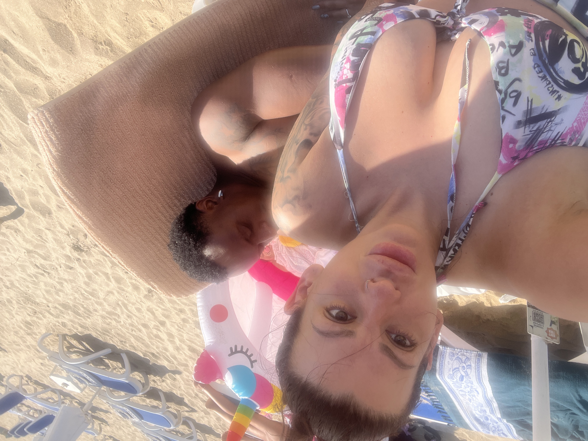 Two individuals, a woman and a man, are lying on a beach in Sozopol, Bulgaria. The woman is in the foreground, facing the camera with her head tilted. She has dark hair pulled up in a bun and is wearing a bikini with a graffiti-like pattern. She is also wearing a nose ring. The man is behind her, lying on his side, with his eyes closed. He has dark curly hair and tattoos on his arms and chest. They are lying on a beach blanket, with an inflatable unicorn float between them. In the background, several beach chairs are visible, some with blue and white stripes. The sand is light brown, and the lighting suggests it is daytime.