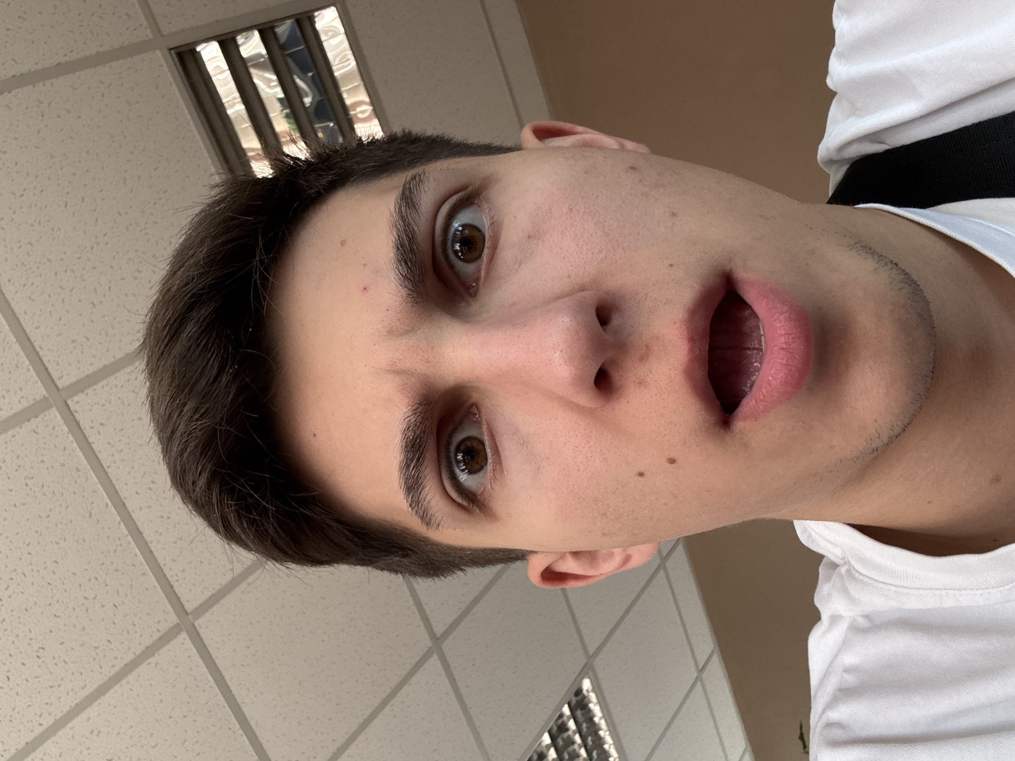 A close-up, low-angle, selfie-style portrait of a young male with dark hair and brown eyes is captured. He has an astonished facial expression, with wide eyes and an open mouth. The background consists of a suspended ceiling with fluorescent lights and a wall. The scene appears to be indoors, possibly in a school or office environment. The individual is wearing a white t-shirt. There is no information in the image to suggest the location is in the city of Shumen, Bulgaria.