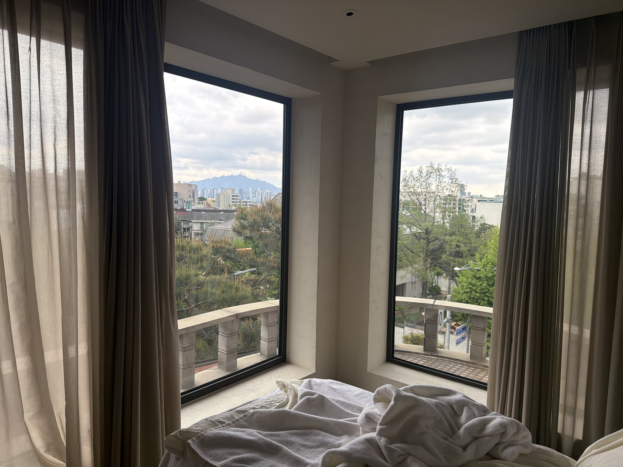 The image displays an interior room, likely a bedroom or hotel room in Seoul, South Korea, featuring two large, floor-to-ceiling windows set into a corner. On either side of the windows, light-colored sheer curtains and darker, heavier drapes are pulled back, allowing light to enter.
Below the windows, a bed is visible with white, rumpled bedding, suggesting recent use or a comfortable, relaxed environment. Just outside the windows, a stone balustrade or railing can be seen, indicating a balcony or a decorative exterior feature.
The outdoor scene, viewed through the windows, depicts an urban landscape under an overcast sky. In the mid-ground, lush green trees and foliage are prominent. Further in the distance, a dense array of city buildings stretches across the horizon, with a distinct mountain range rising majestically behind them. A blue street sign, partially visible through the right window, displays Korean characters, confirming the location as Seoul. The time of day appears to be daytime, likely cloudy given the soft, diffused light. The overall mood is peaceful and comfortable, offering a scenic urban view from an intimate indoor setting.